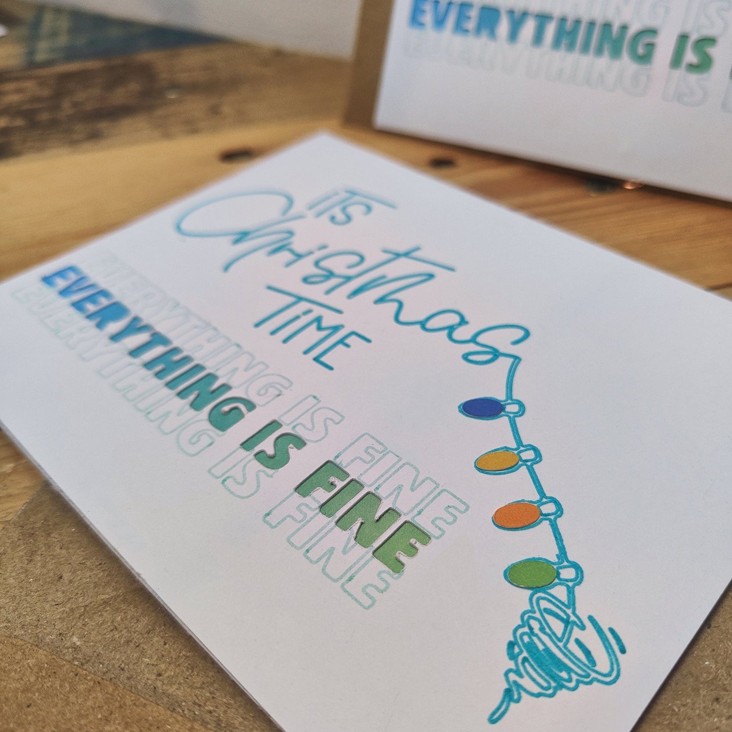 Card with 'It's Christmas Time, Everything is Fine' text and Christmas lights design getting pulled into a tornado on a wooden surface.