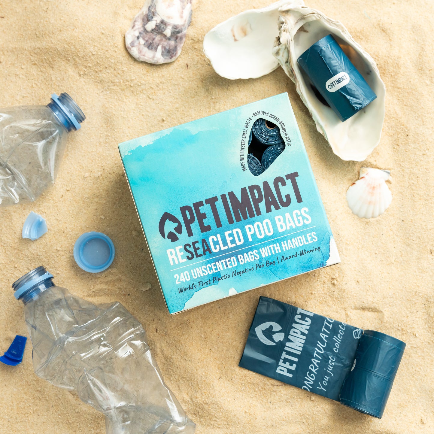 Pet Impact ReSEAcled dog poo bags with packaging, individual roll and oyster shell displayed on a sandy beach with recycled plastic bottles to highlight the product's recycled materials.