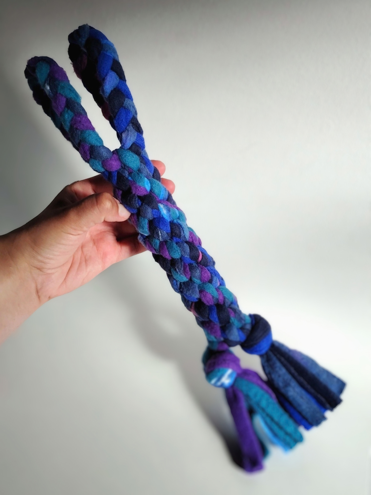 Hand holding a Nova Zeal Night Sky fleece tug toy, showing its braided construction in navy, royal blue, teal and purple, with knotted tassels at the end.
