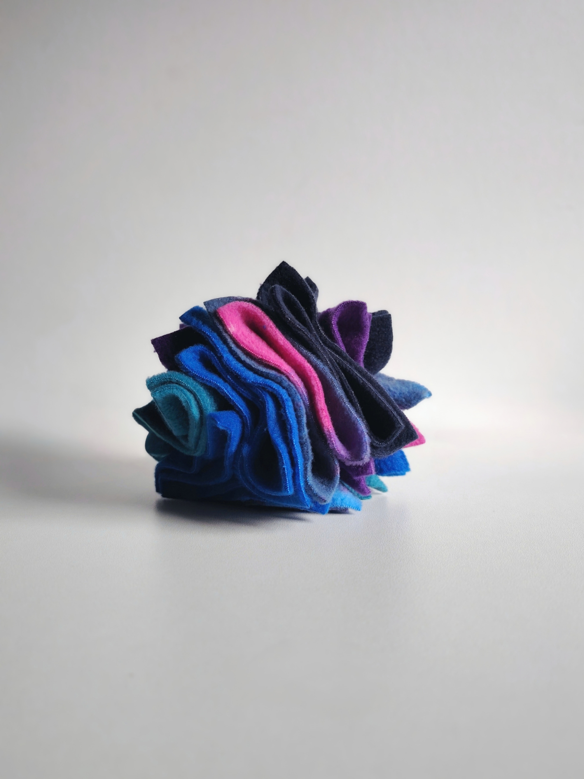 Side view of the Nova Zeal Night Sky Small Scruffle Ball, showing layered folds of navy, royal blue, teal, purple and galaxy-patterned fleece against a pale background.