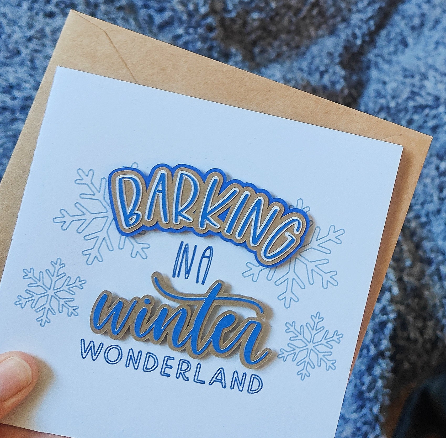 Greeting card with 'Barking in a Winter Wonderland' text on a white background with blue snowflakes, held over a textured blue fabric.