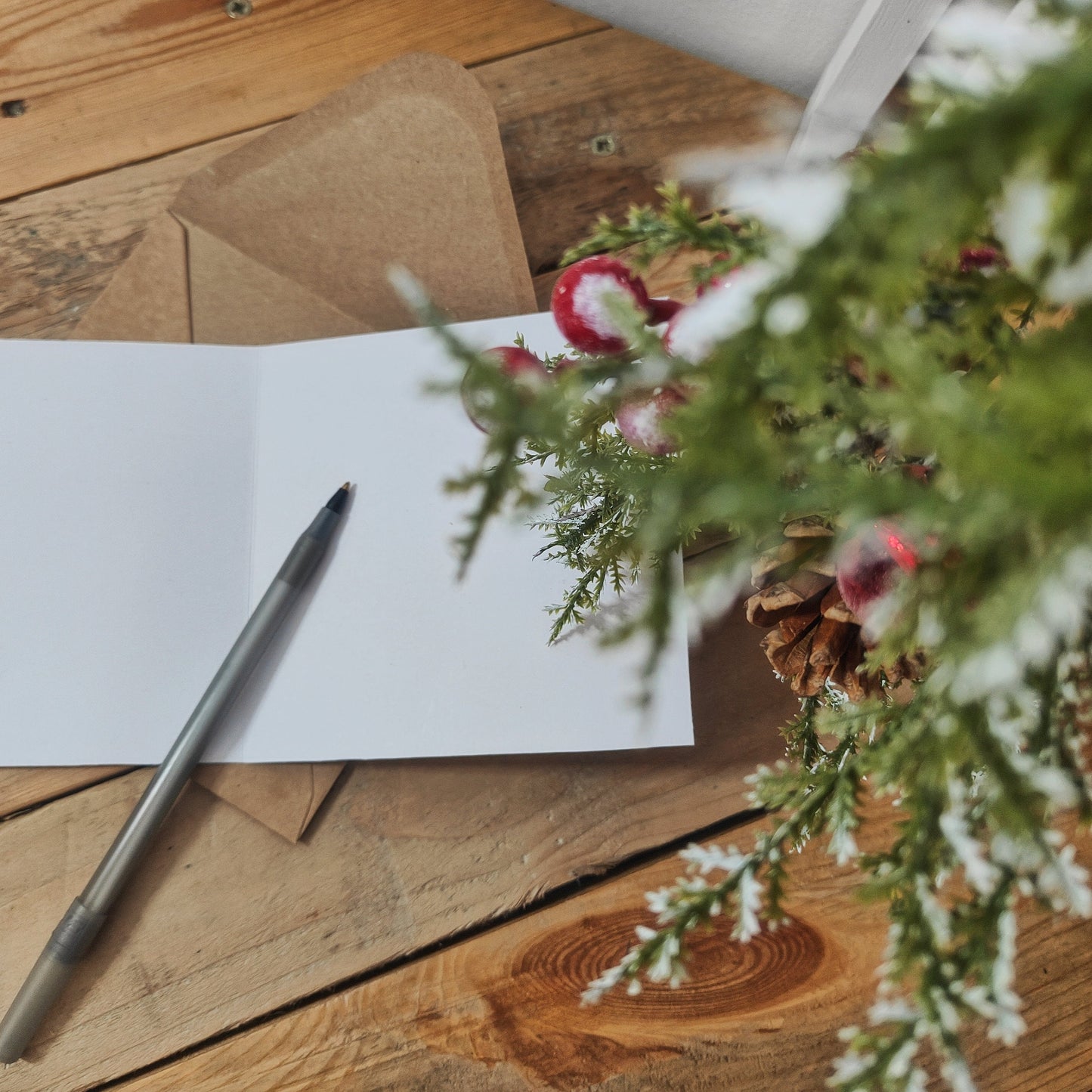 Open Christmas card with pen and envelope on wooden surface with decorative plants.
