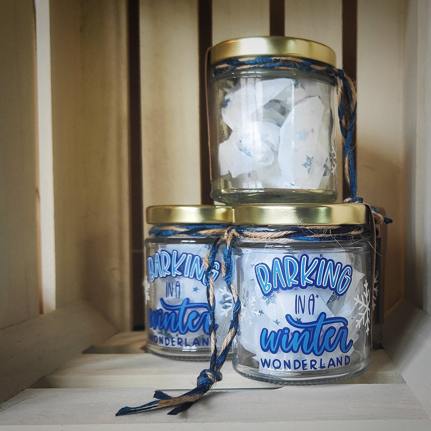 Three glass jars with gold lids and blue string/jute, labeled 'Barking in a Winter Wonderland', stacked on a wooden surface. The jar stacked on top is showing the clear glass on the reverse of the jar.