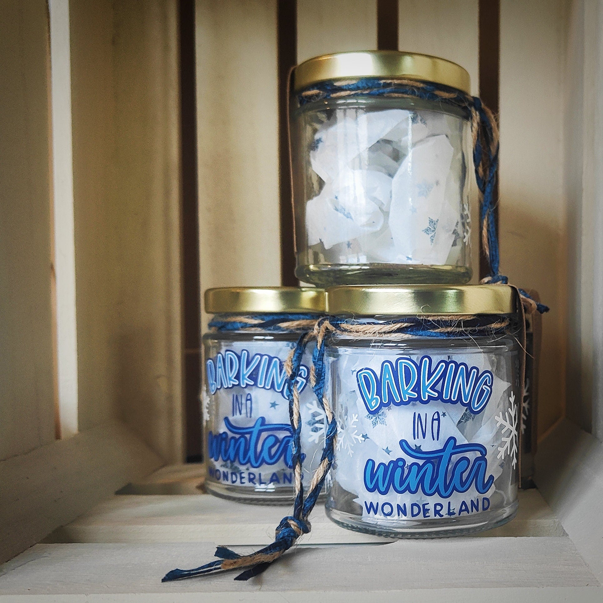 Three glass jars with gold lids and blue string/jute, labeled 'Barking in a Winter Wonderland', stacked on a wooden surface. The jar stacked on top is showing the clear glass on the reverse of the jar.