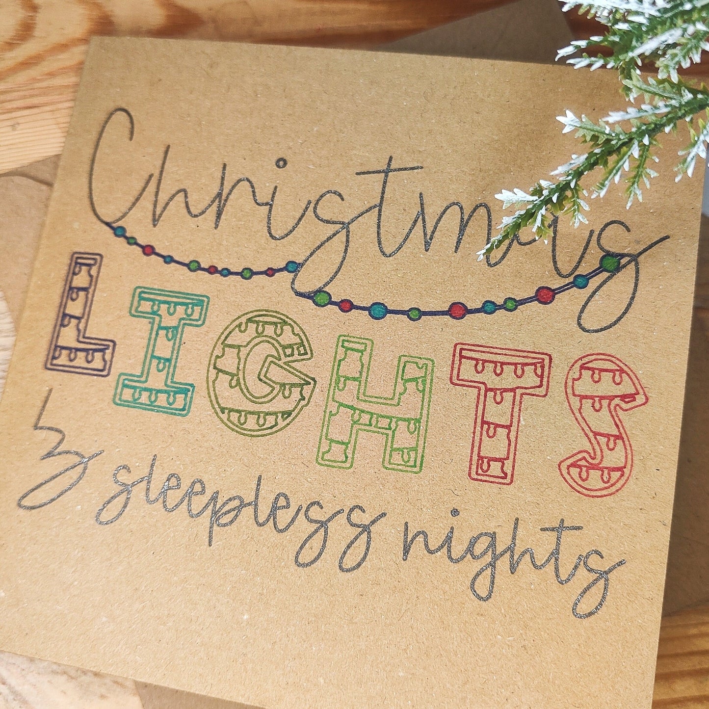 Card with 'Christmas Lights & Sleepless Nights' text on a wooden surface with winter greenery.