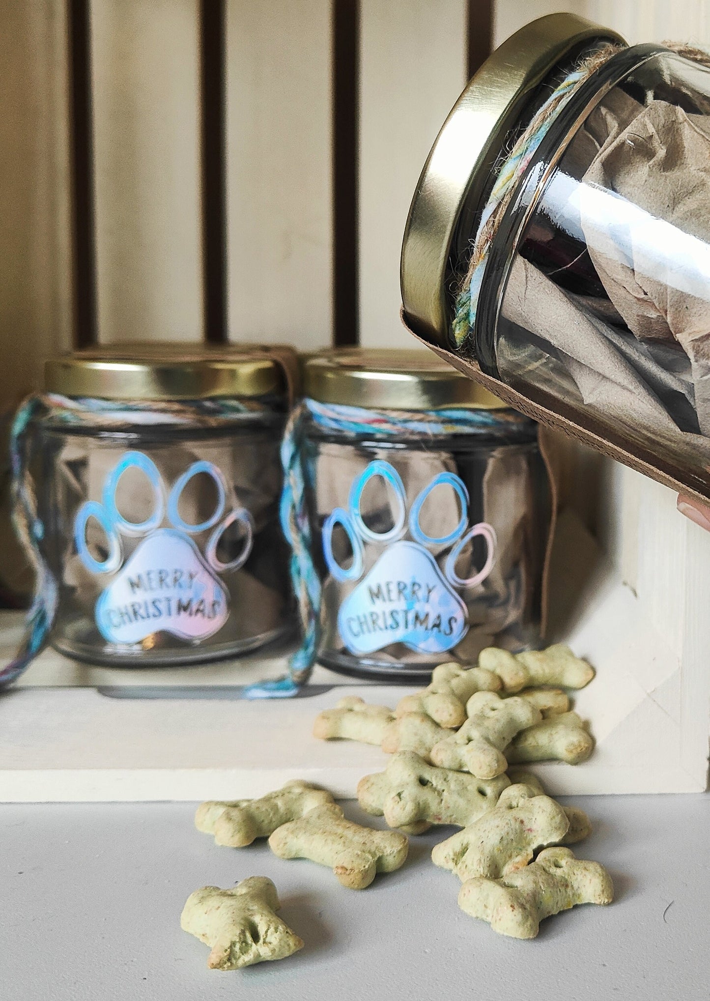 Two glass jars with paw print designs and 'Merry Christmas' text, surrounded by dog treats on a surface.