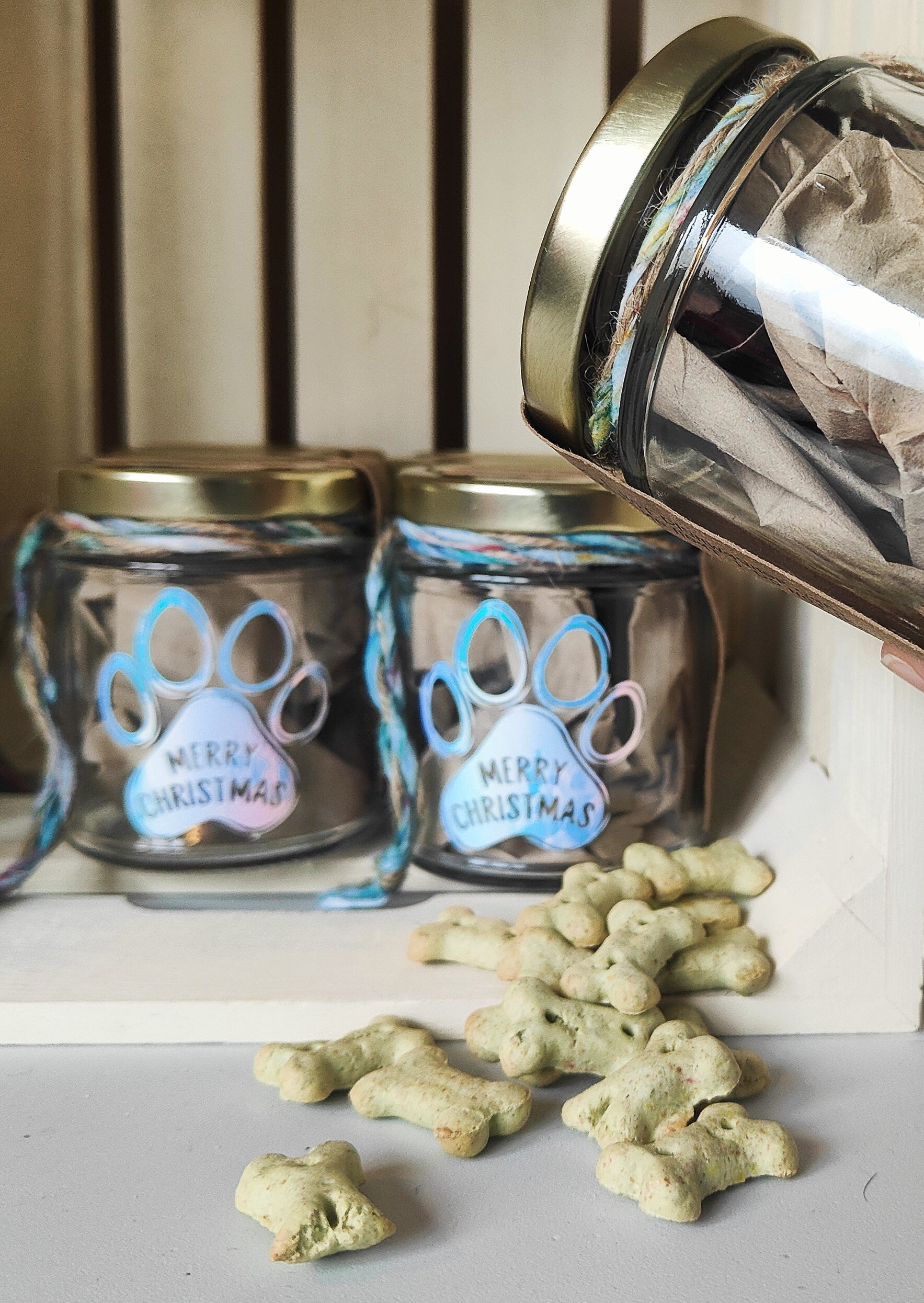 Two glass jars with paw print designs and 'Merry Christmas' text, surrounded by dog treats on a surface.