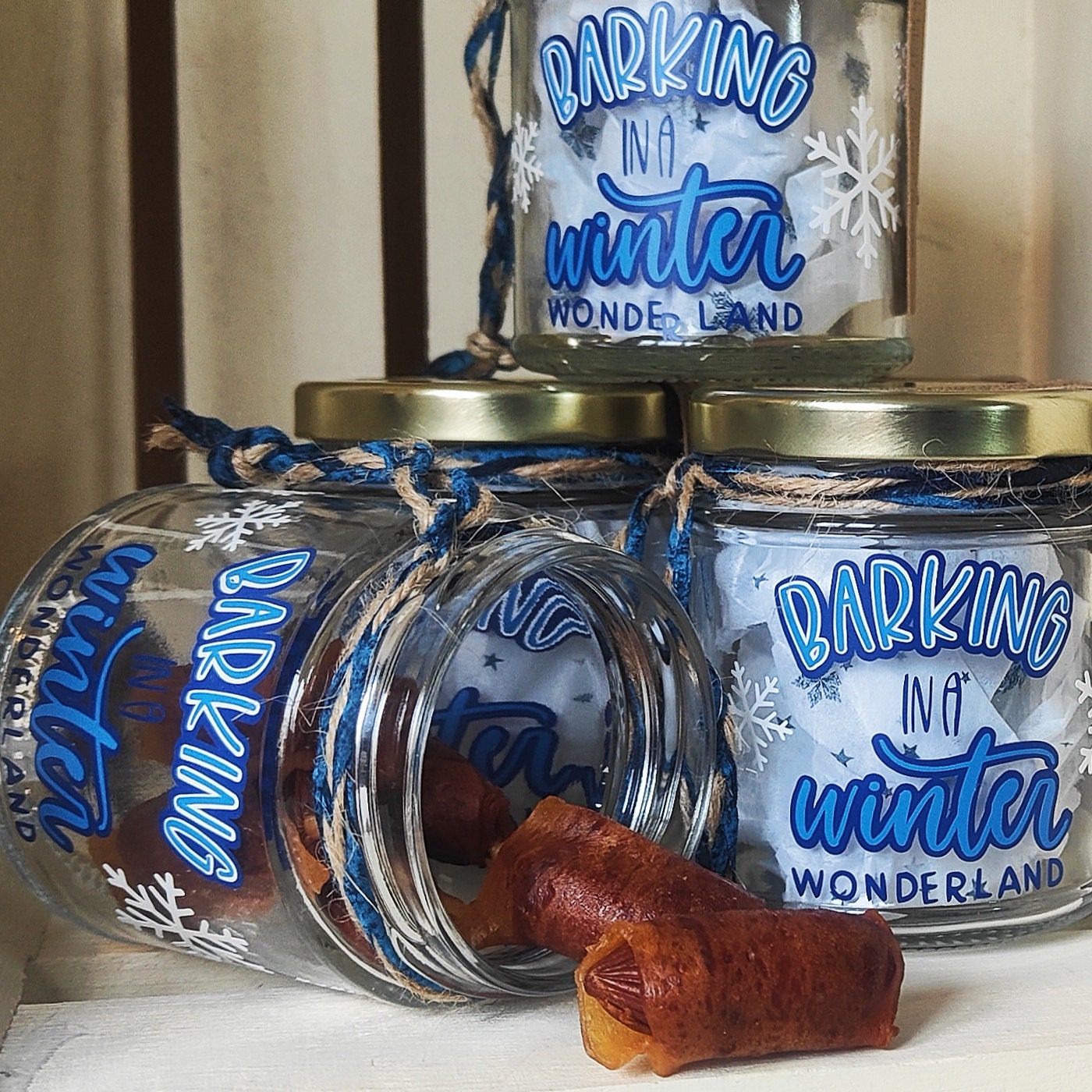 Glass jars with 'Barking in a Winter Wonderland' text, tied with blue string/jute, on a light background. Dog Treats Spilling out of one jar as an example.
