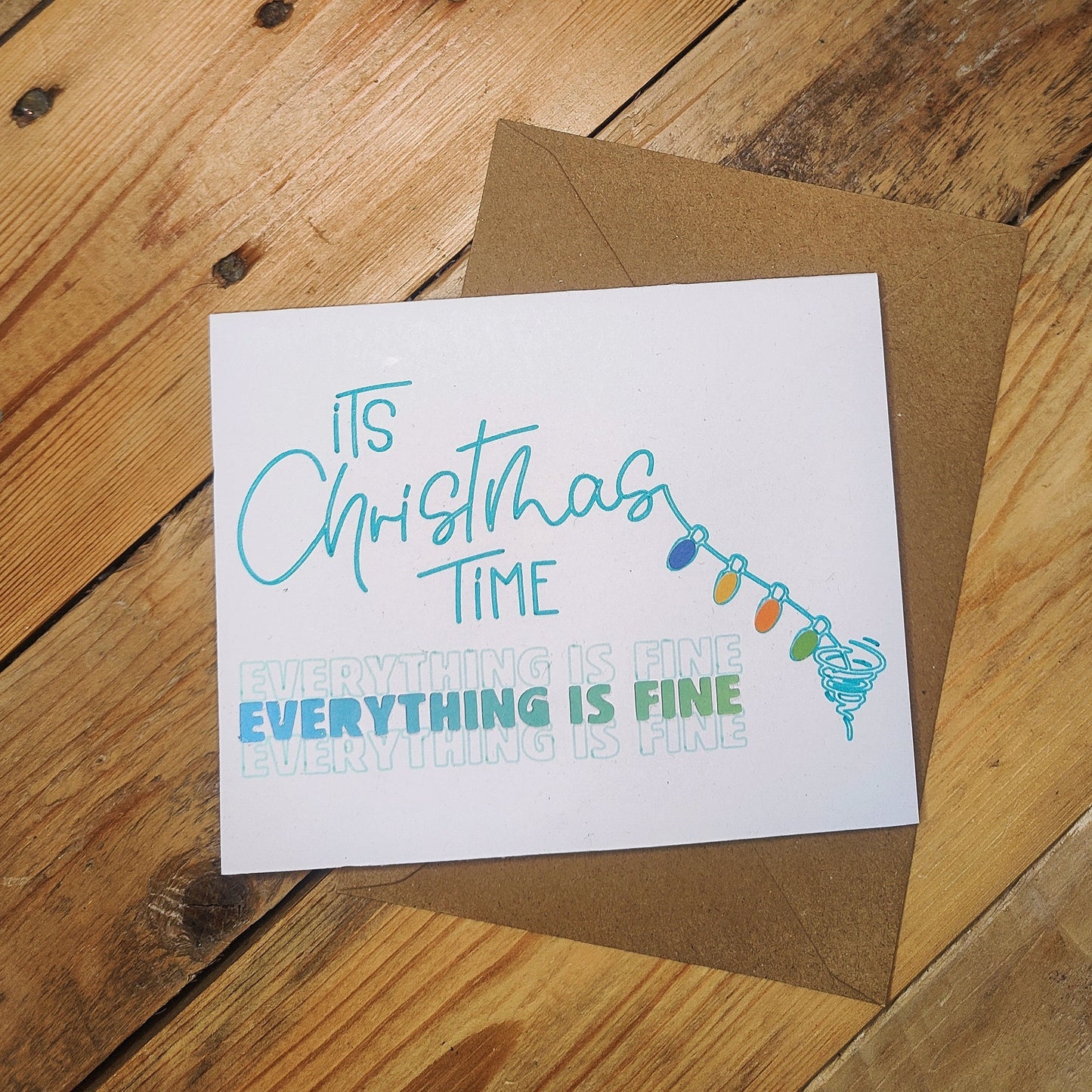 Christmas-themed card with text saying ' It's Christmas TIME... EVERYTHING IS FINE' and string lights on a wooden surface