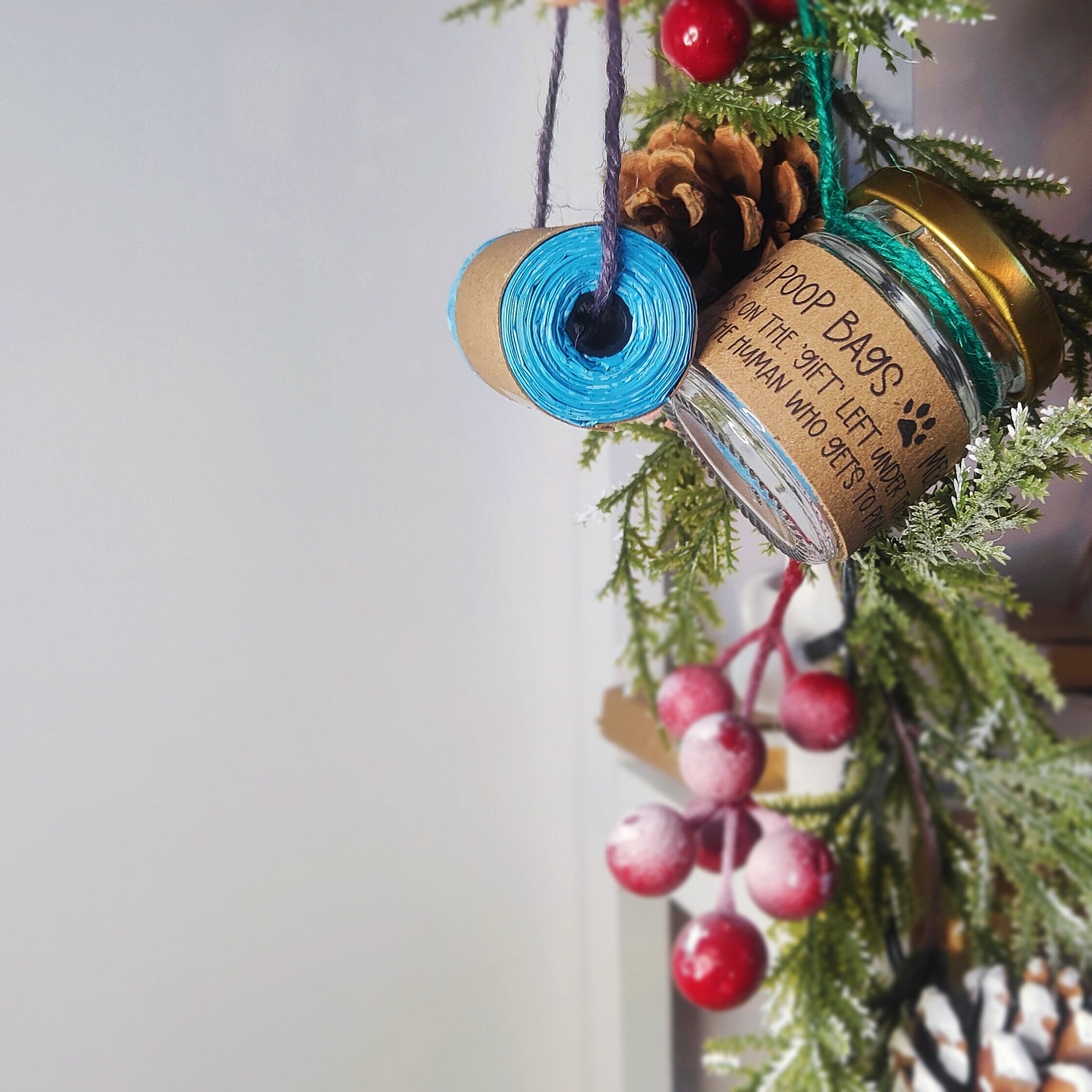 A handmade Christmas tree decoration featuring a jar with a Christmas-themed label, wrapped in jute twine, and hanging from a Christmas tree decoration. The label includes a humorous message about 'emergency poop bags' for dogs.