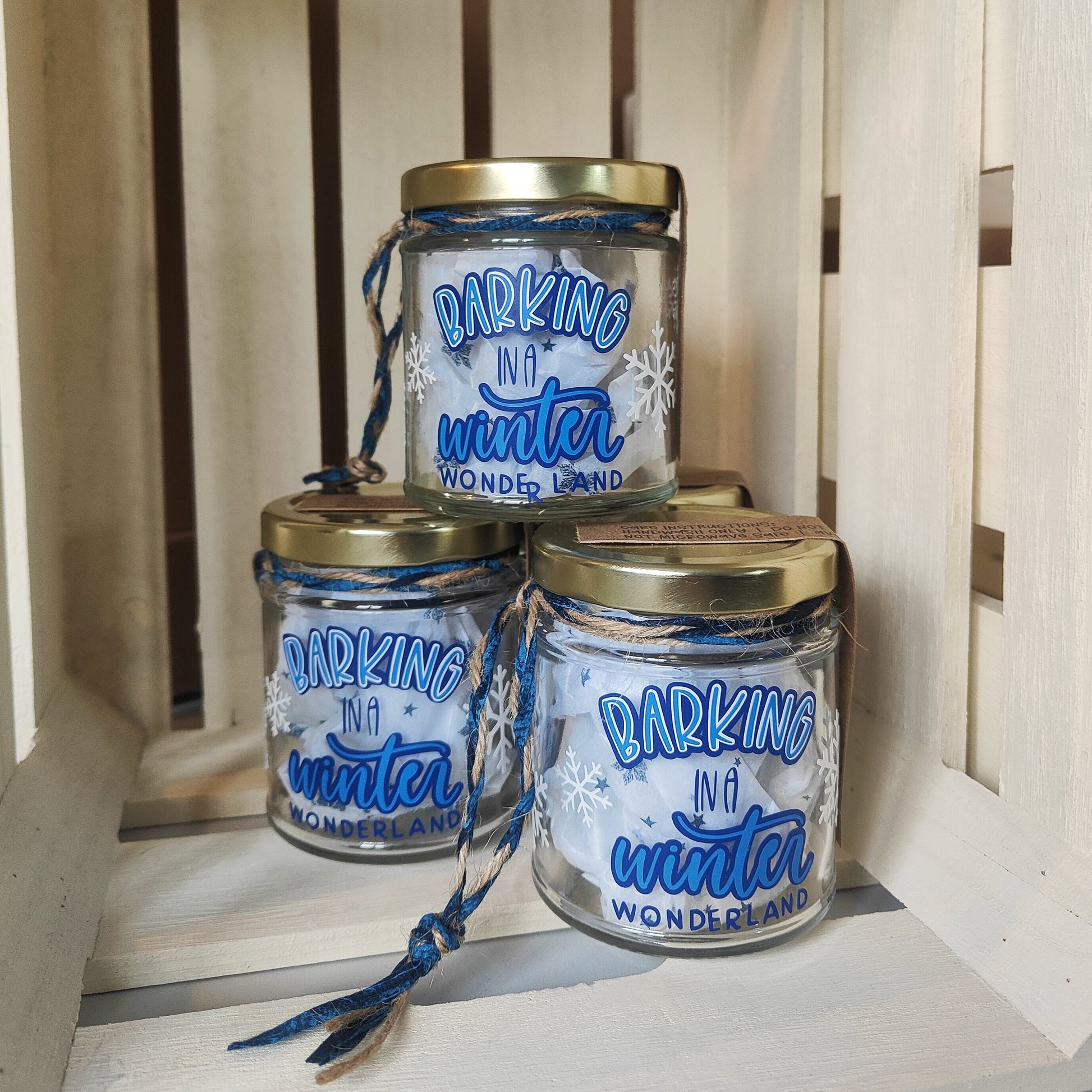 Three jars with gold lids and blue text 'Barking in a Winter Wonderland' on a wooden surface. Filled with tissue paper to show they are empty.