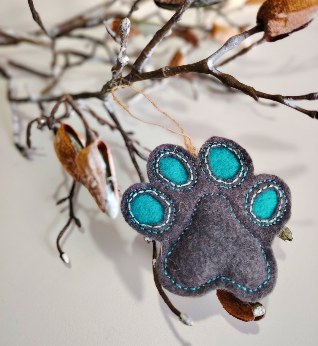 Grey Felt paw print ornament with teal centers hanging on a branch.