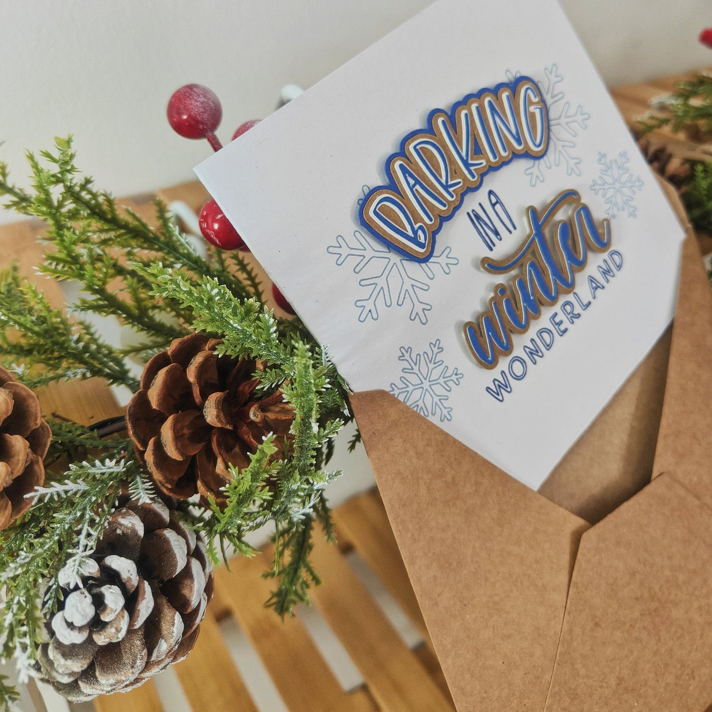 Greetings card halfway inside a kraft envelope, surrounded by pine cones and greenery.