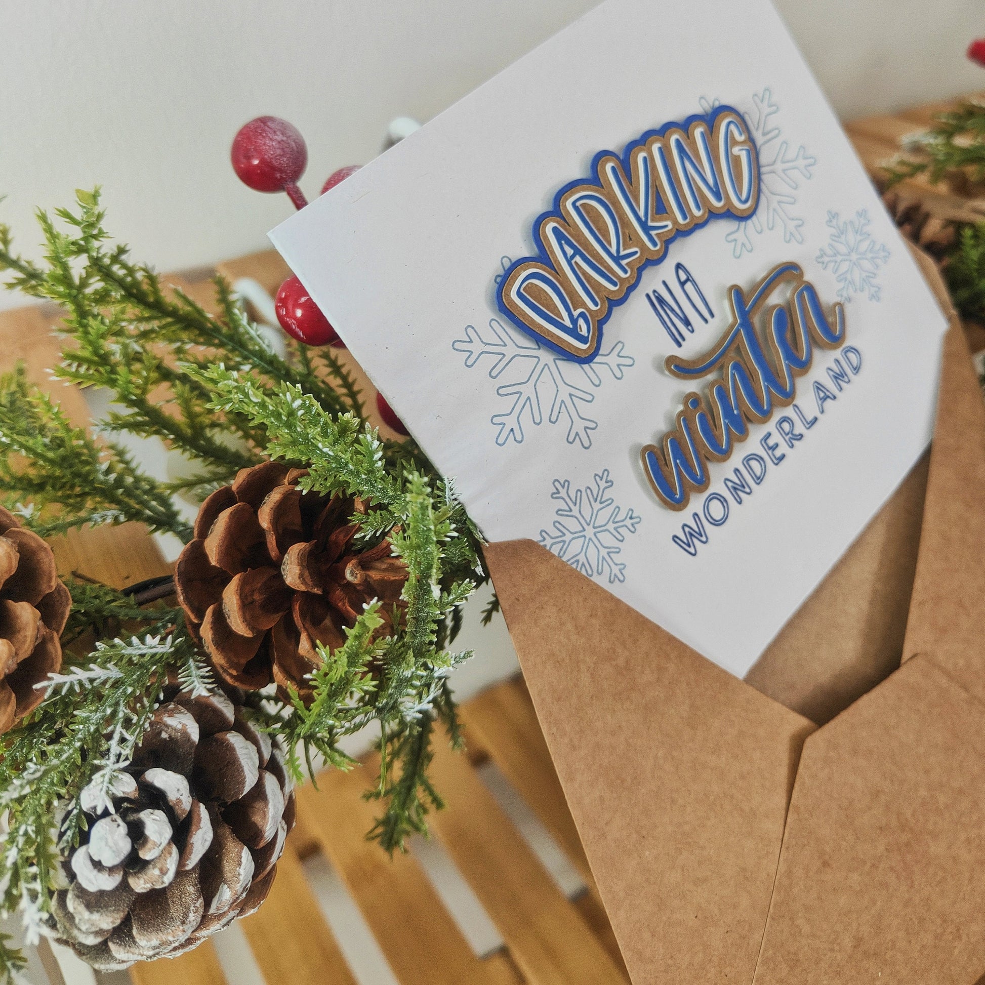 Greetings card halfway inside a kraft envelope, surrounded by pine cones and greenery.