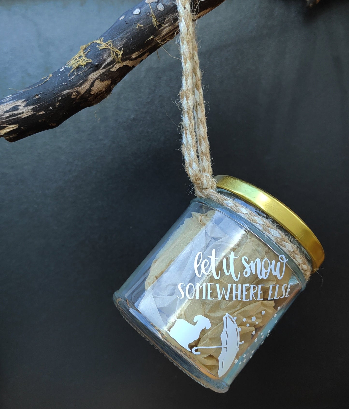 Decorative jar with 'let it snow somewhere else' text and a gold lid, hanging on a branch.