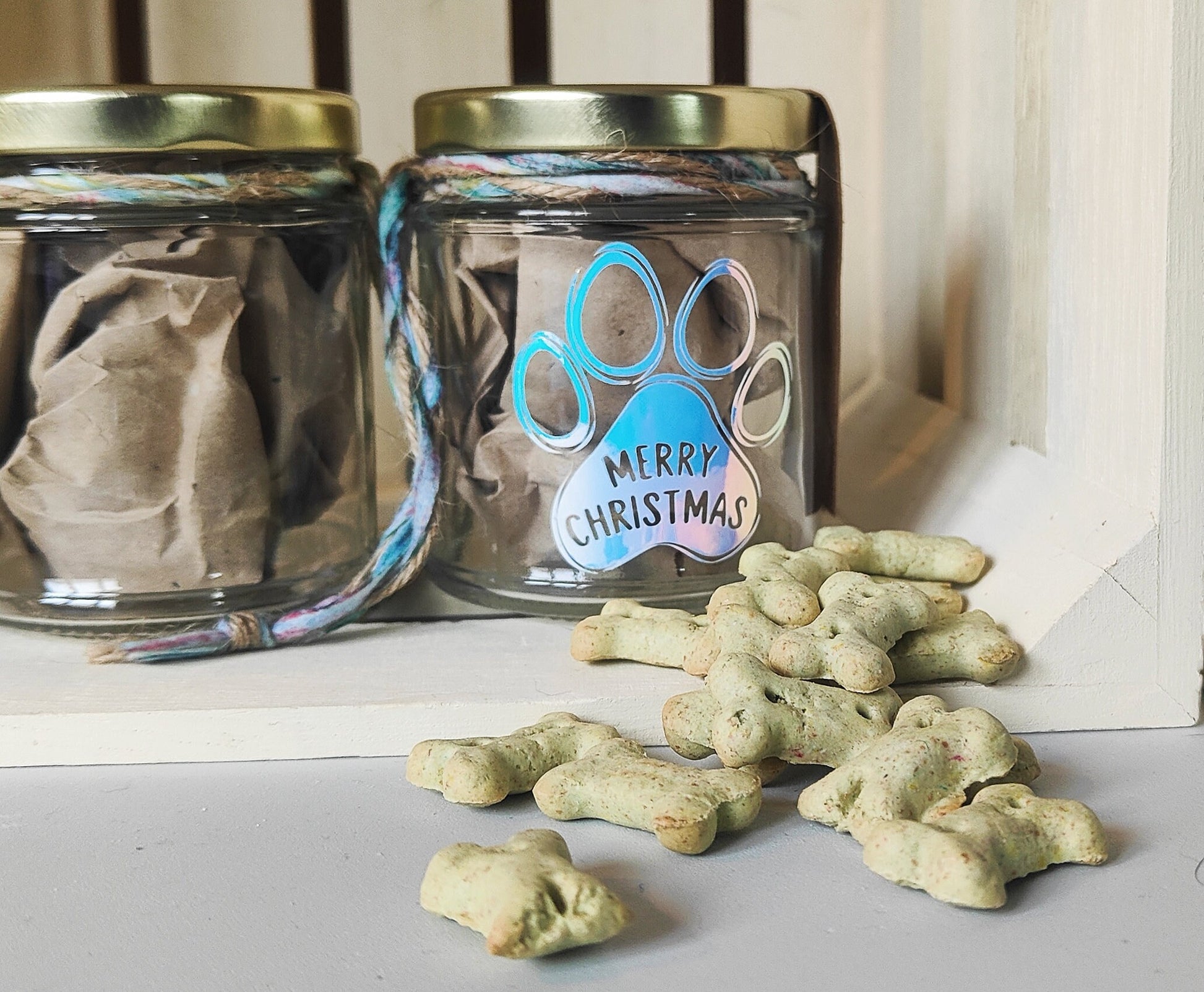 Two glass jars with gold lids and a paw print design, accompanied by dog treats on a white surface.