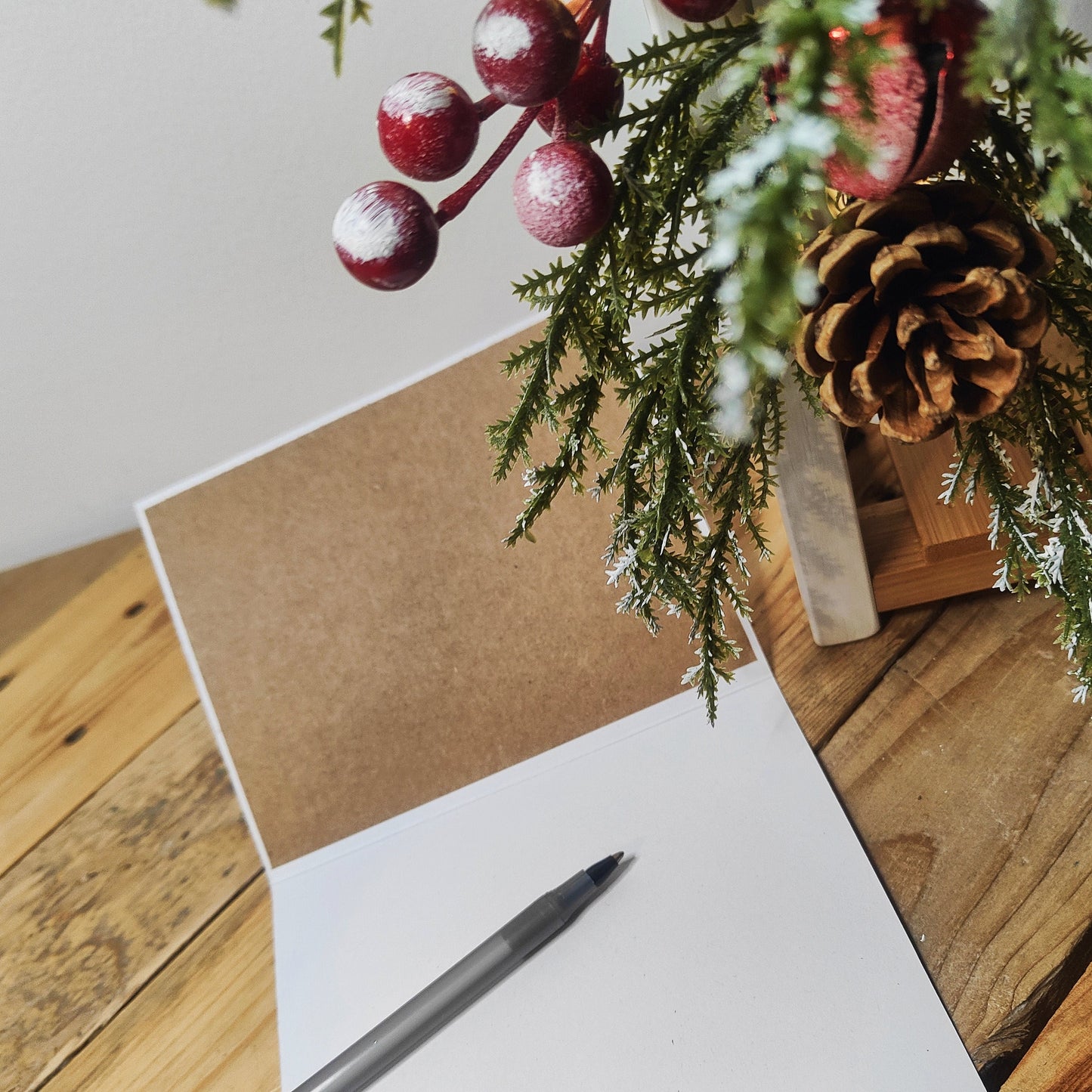 White greeting card showing one side is kraft brown card inside, pen, and Christmas decorations on a wooden surface