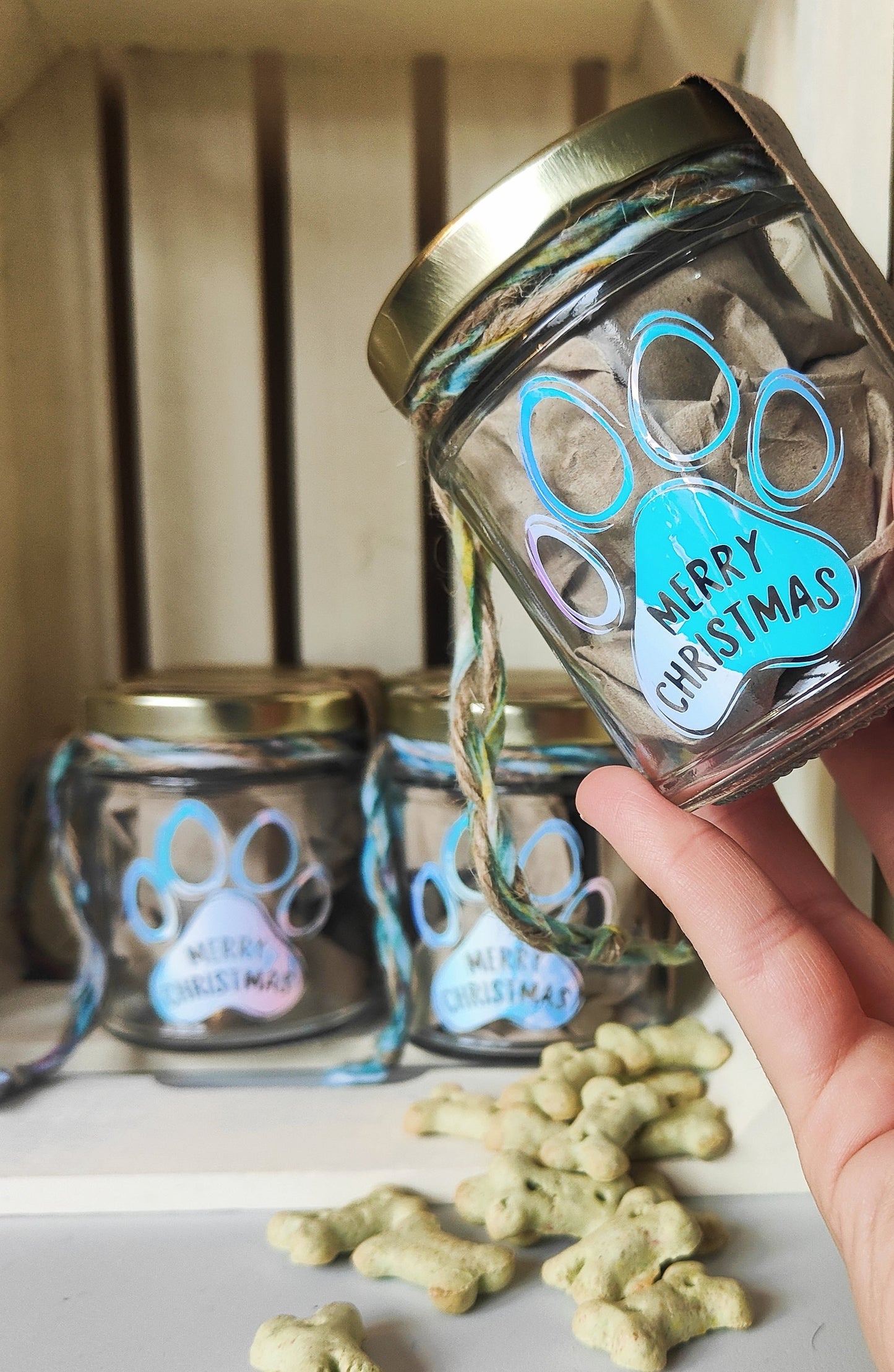 Glass jar with gold lid and paw print design, held by a hand with treats on a surface.
