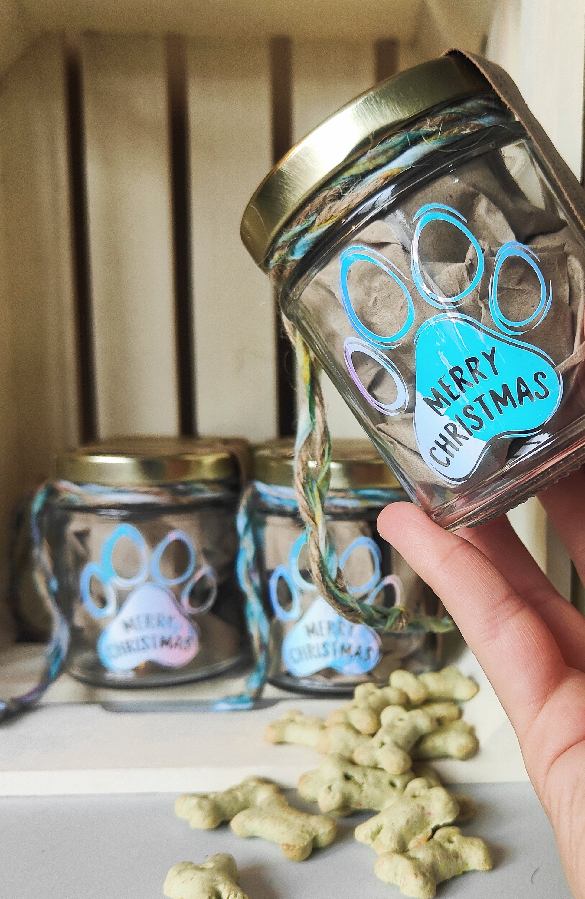 Glass jar with gold lid and paw print design, held by a hand with treats on a surface.