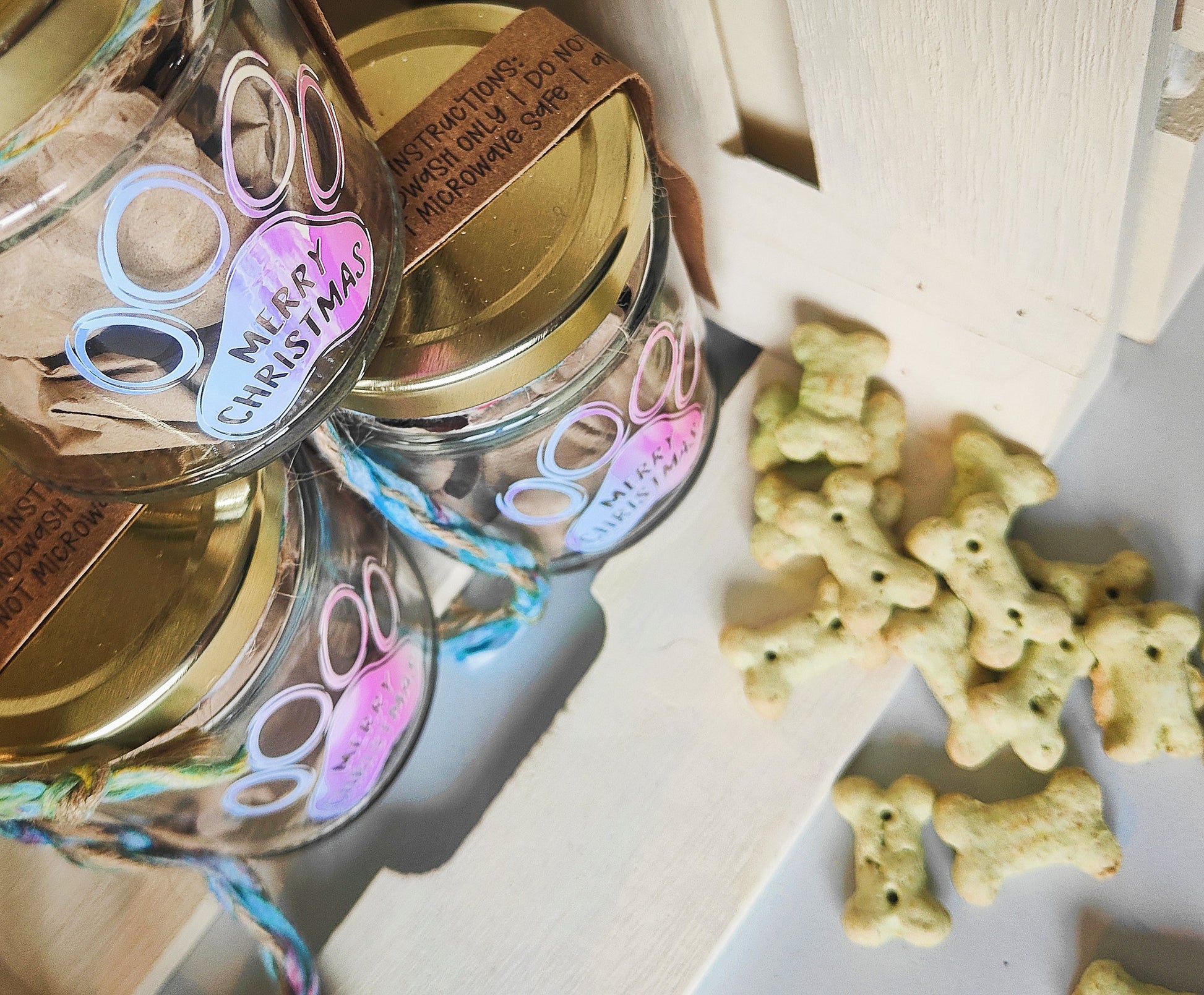 Glass jars with gold lids, labeled 'Merry Christmas', next to dog treats on a white surface.