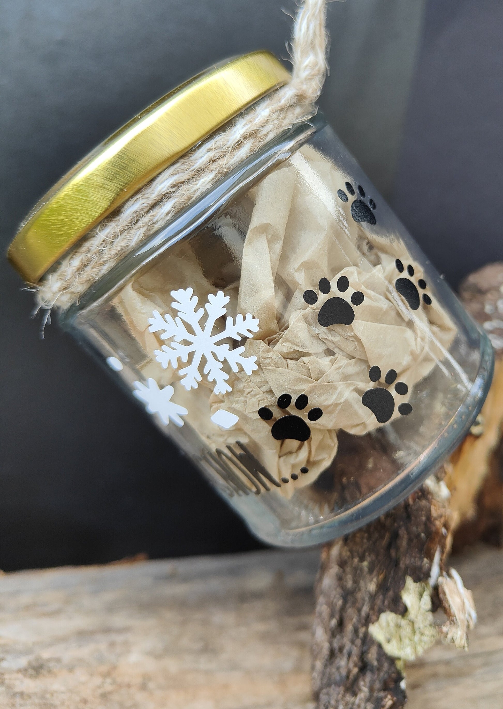 Glass jar hanging from natural jute with gold lid, paw prints, and snowflakes, with a wooden surface behind.