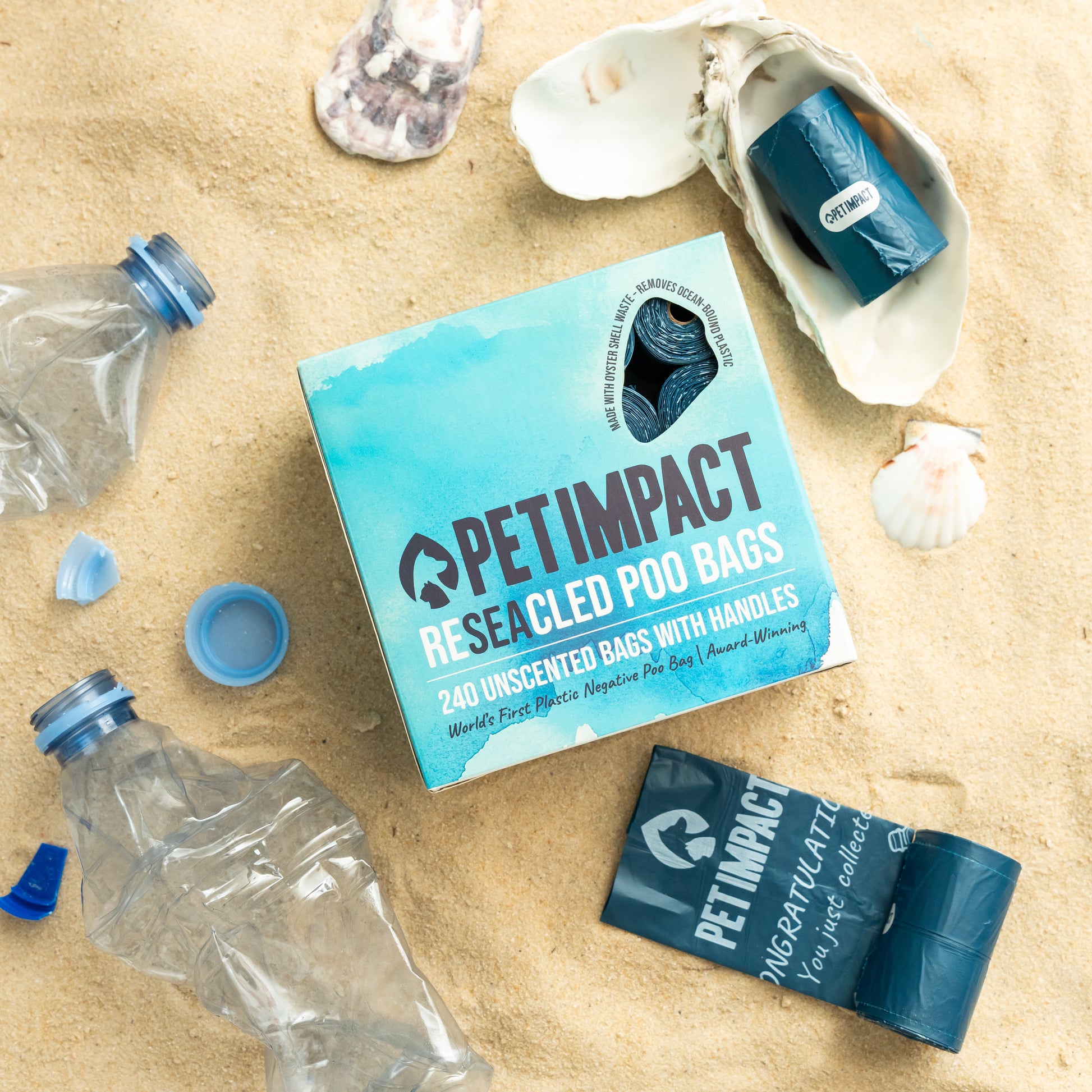 Pet Impact ReSEAcled dog poo bags with packaging, individual roll and oyster shell displayed on a sandy beach with recycled plastic bottles to highlight the product's recycled materials.