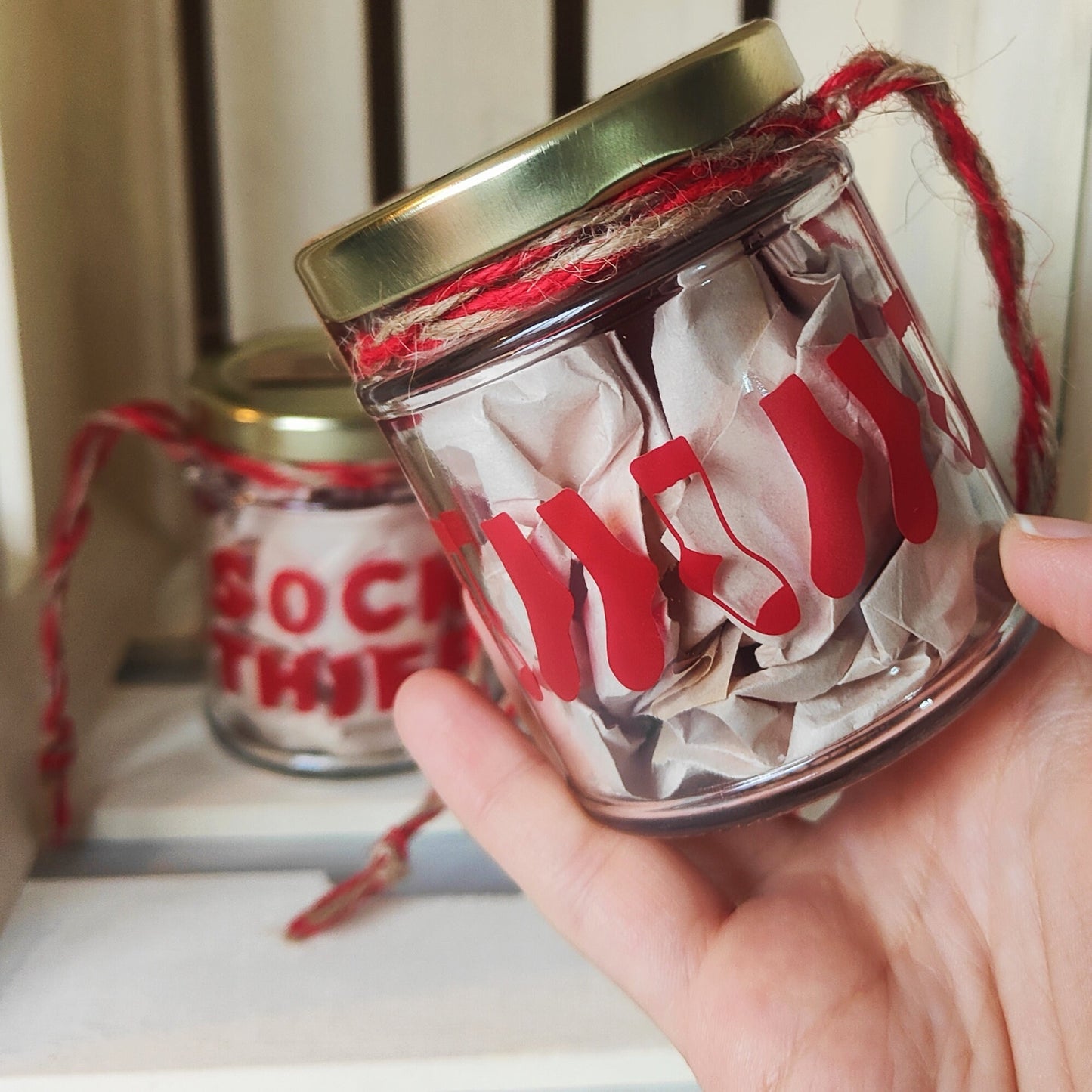 Hand holding a glass jar with red socks on the back of the 'Sock Thief' Jar and a gold lid, with another jar in the background showing the front of the design.