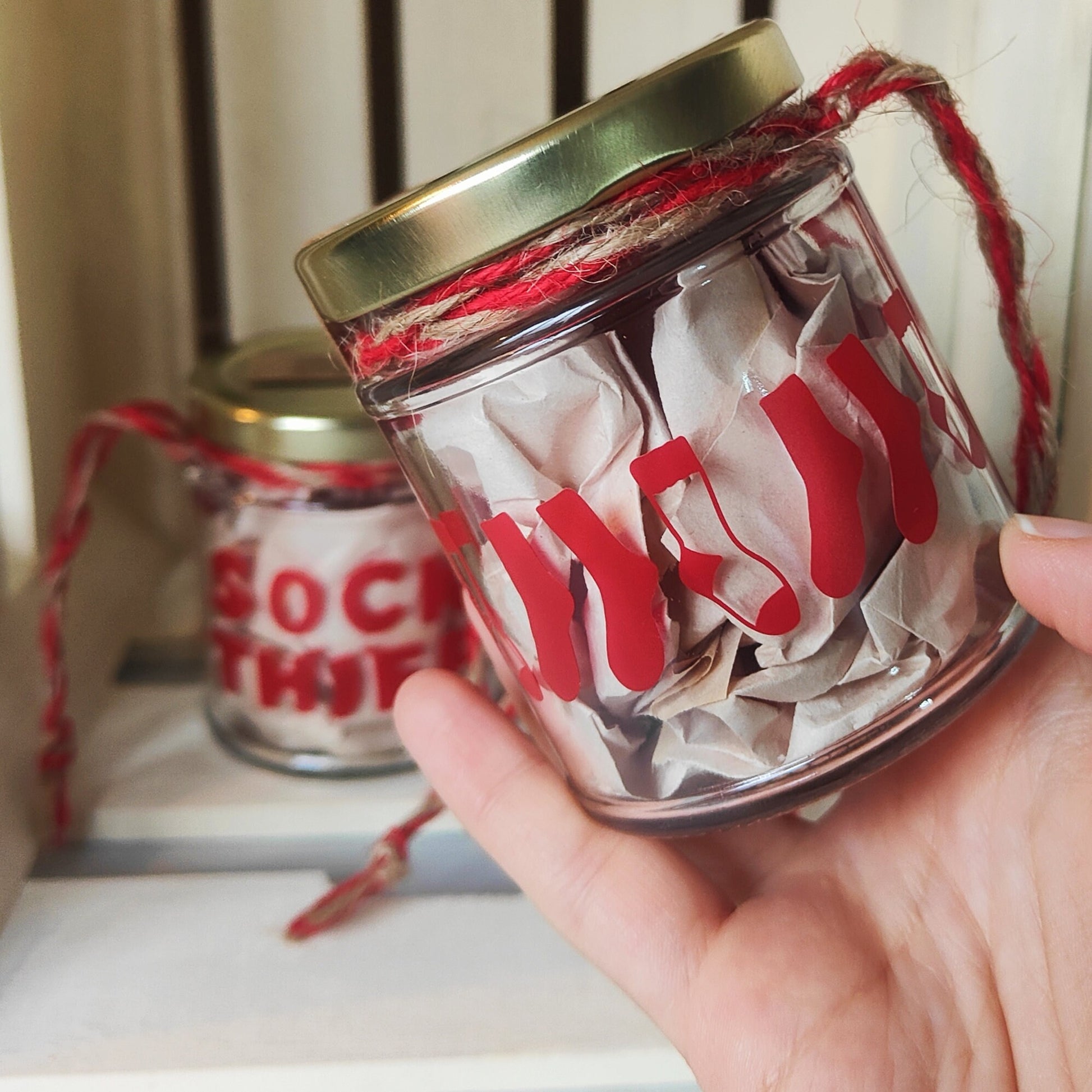 Hand holding a glass jar with red socks on the back of the 'Sock Thief' Jar and a gold lid, with another jar in the background showing the front of the design.