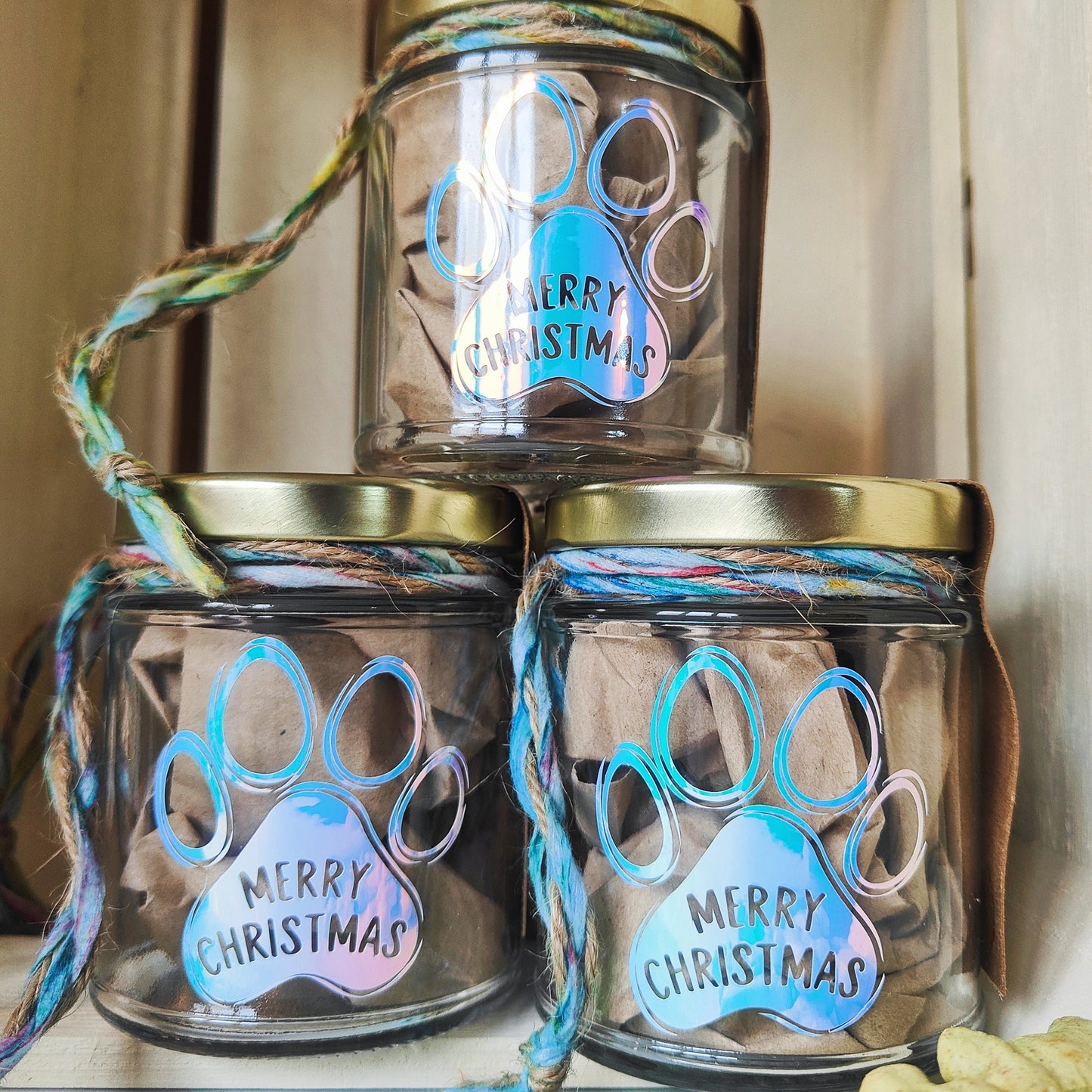 Three glass jars with gold lids, decorated with a paw print and 'Merry Christmas' text, on a neutral background.
