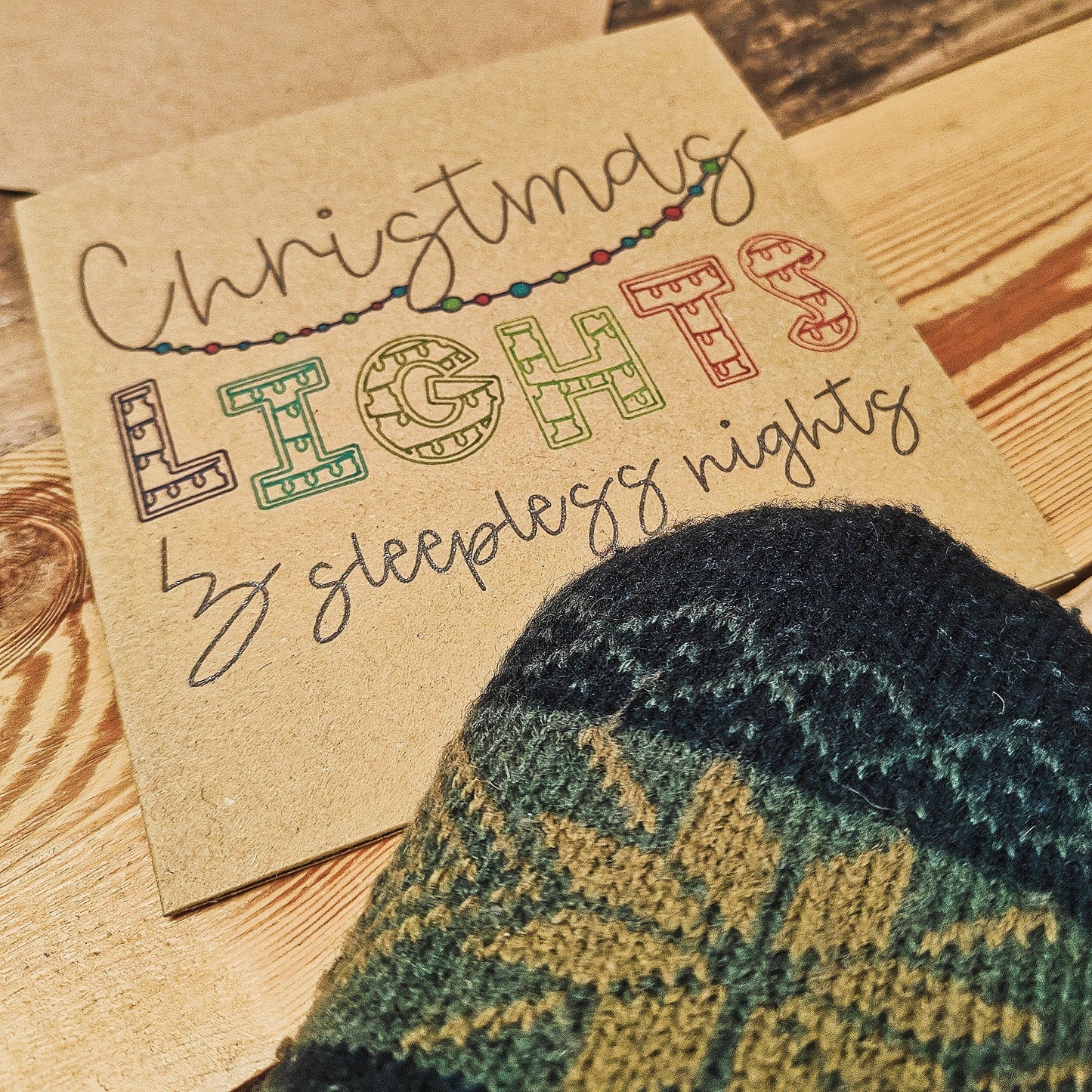 Kraft card with 'Christmas Lights & Sleepless Nights' text and a knitted mitten on a wooden surface