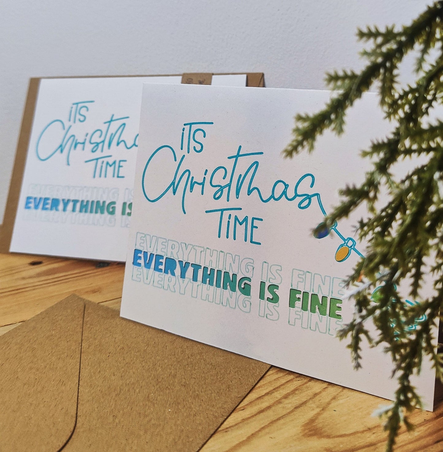 Two Christmas-themed greeting cards on a wooden surface with a branch in the background.