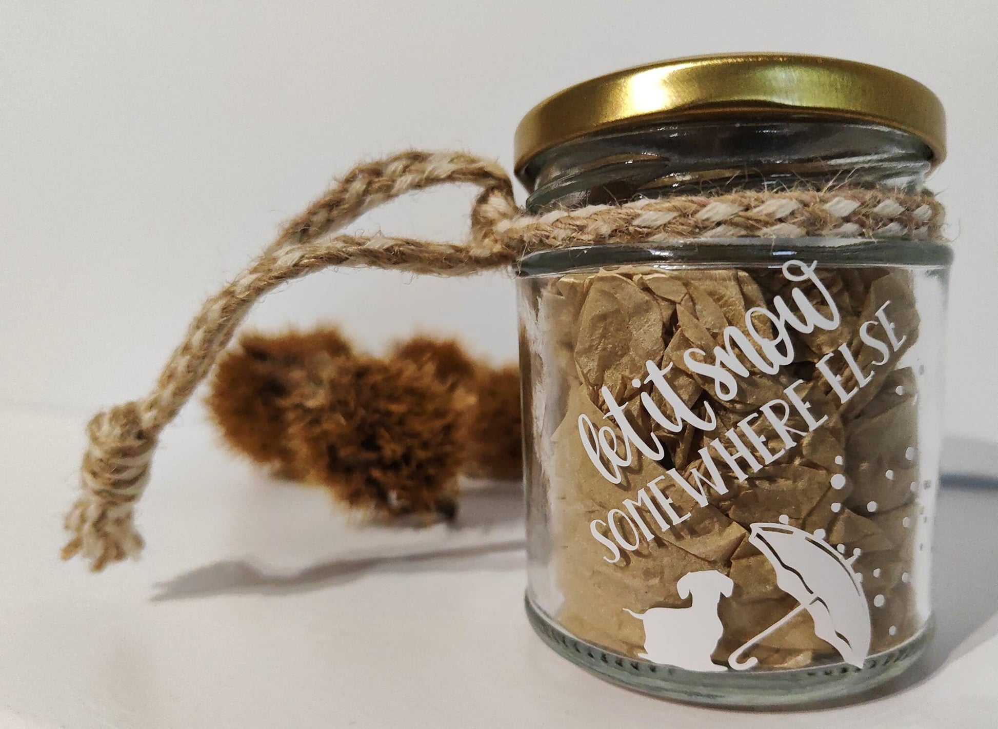 Glass jar with decorative text and a gold lid on a light background. White and natural jute surrounds the neck of the jar for hanging.