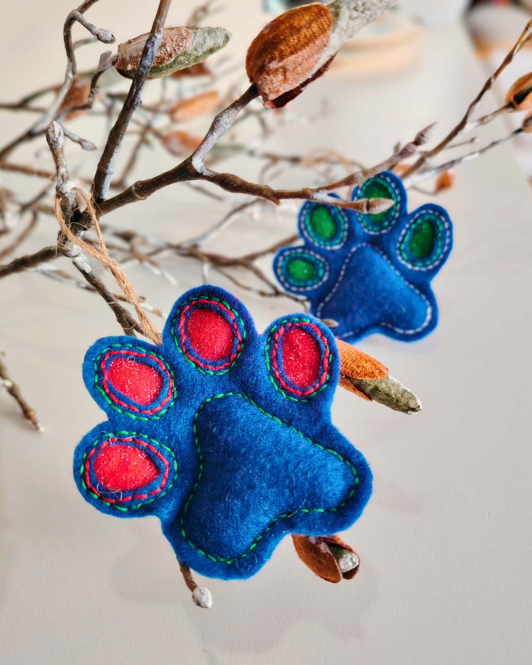 Two Blue paw print decorations one with red, the other with a green variation with a branch blurred in the background
