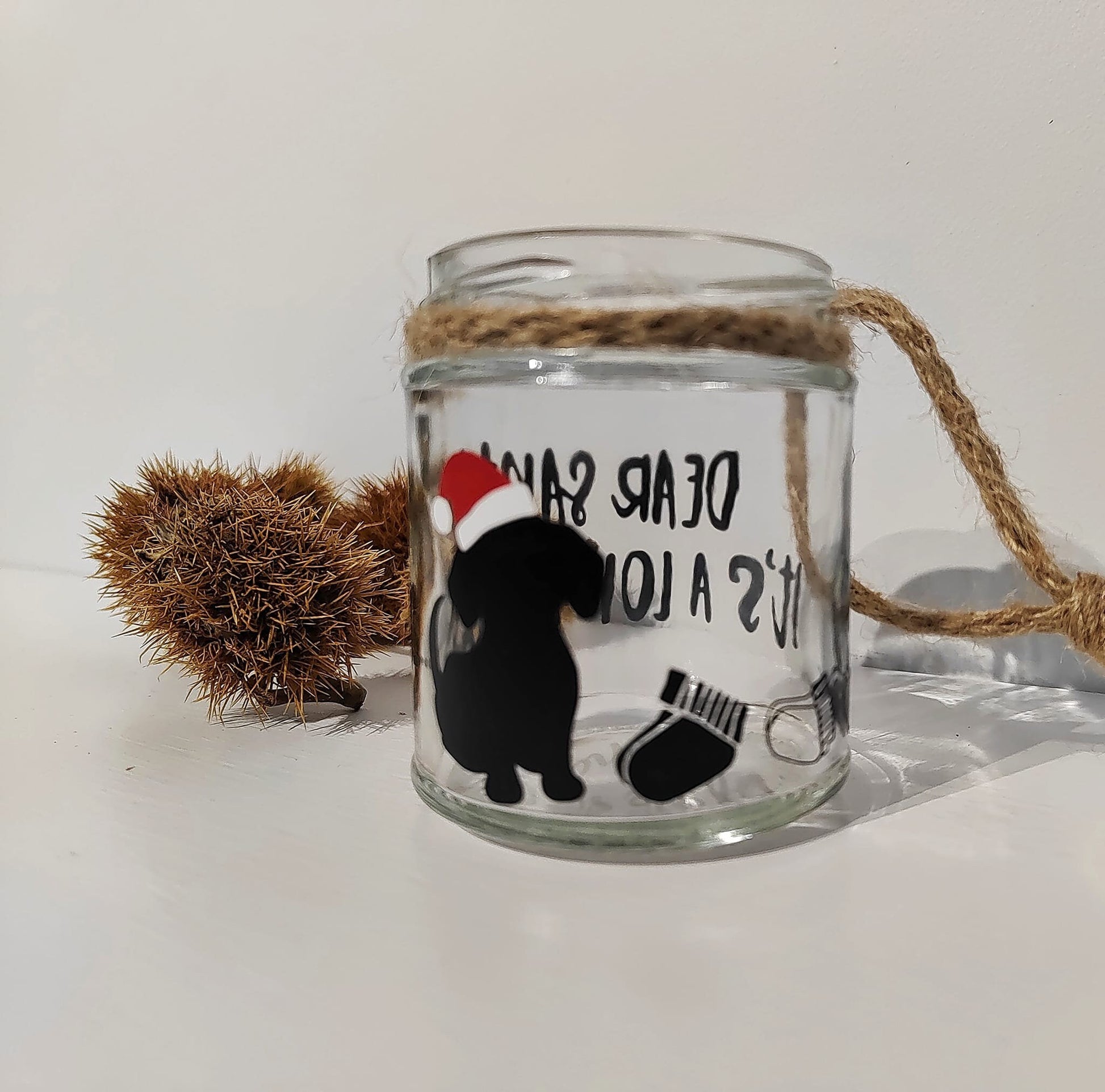 Glass jar with a decorative design of a dog wearing a Santa hat and text, tied with a jute handle on a white background.