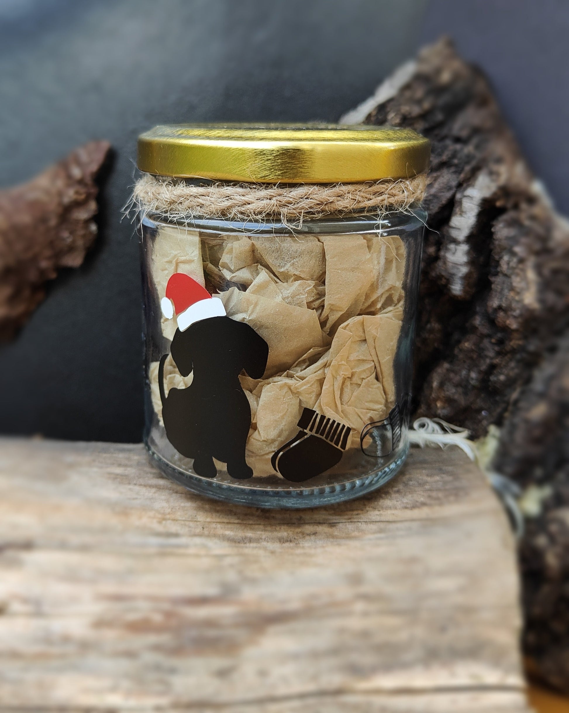 Glass jar with a gold lid featuring a black silhouette of a dog wearing a Santa hat with some socks next to it on a wooden surface