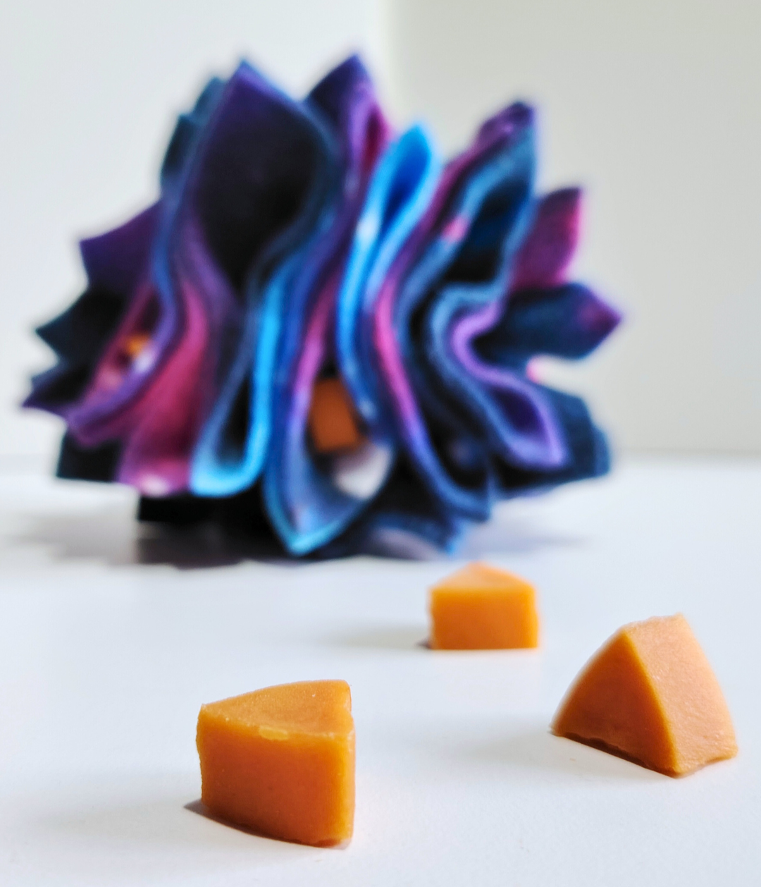 Three orange triangular dog treats on a white surface with a blurred purple and blue snuffle ball in the background.