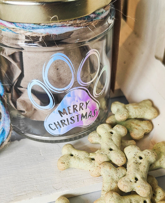 Glass jar with a holographic  paw print design and 'Merry Christmas' text, surrounded by dog treats on a wooden surface.