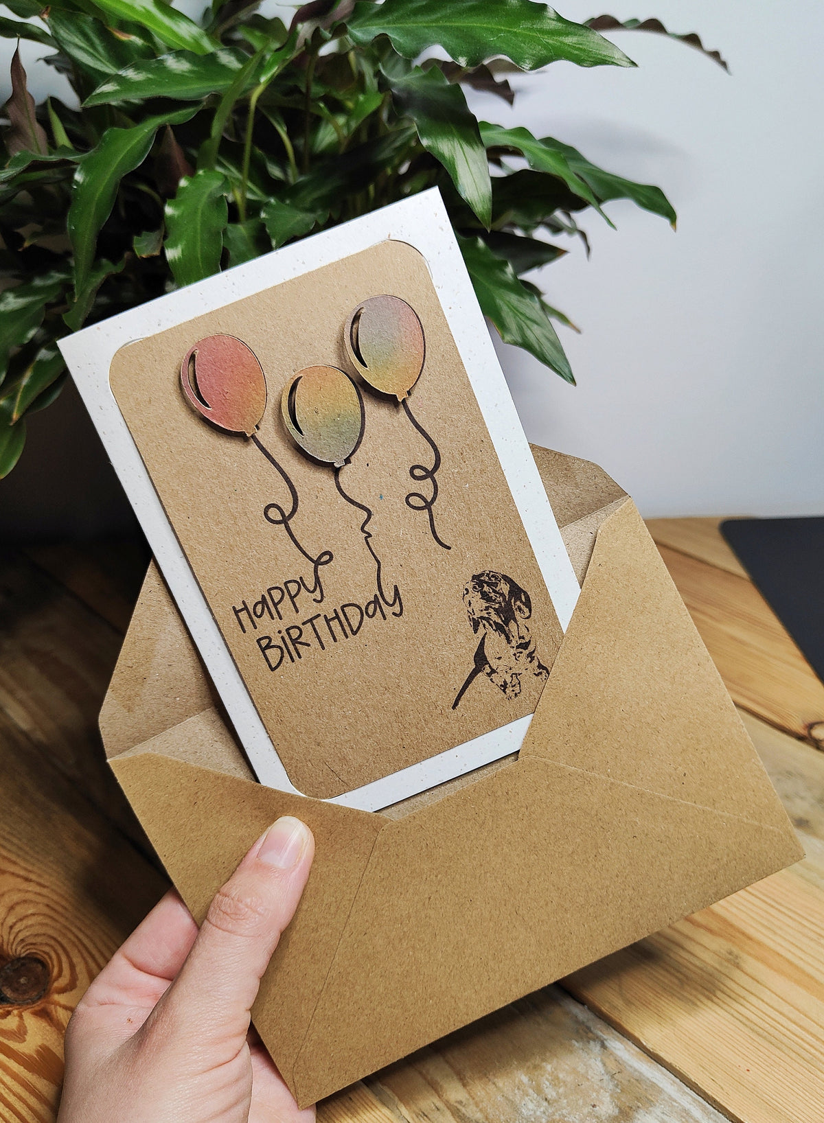 Miniature Dachshund birthday card with colourful balloons, held above a kraft envelope.