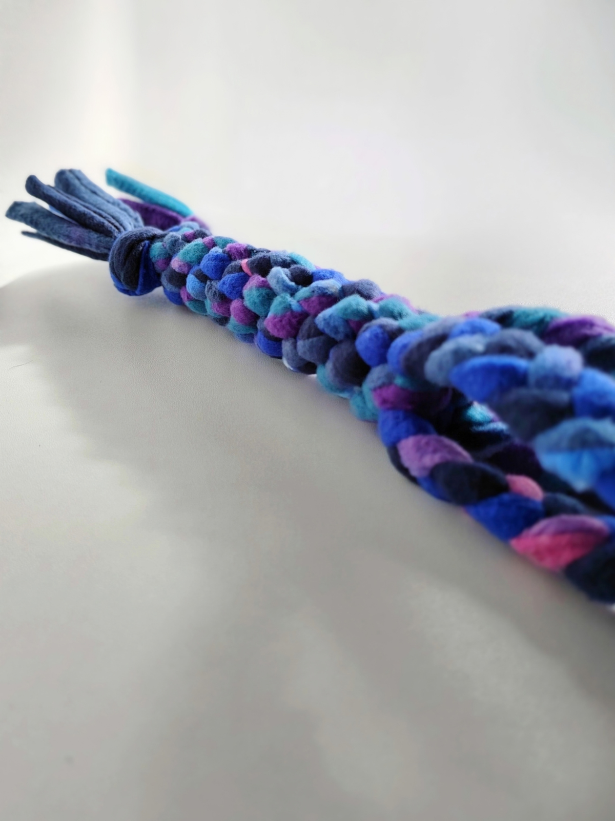 Angled close-up of a handmade Nova Zeal Night Sky handled tug toy lying on a light surface, showing its braided navy, royal blue, teal and purple fleece with a softly blurred foreground.