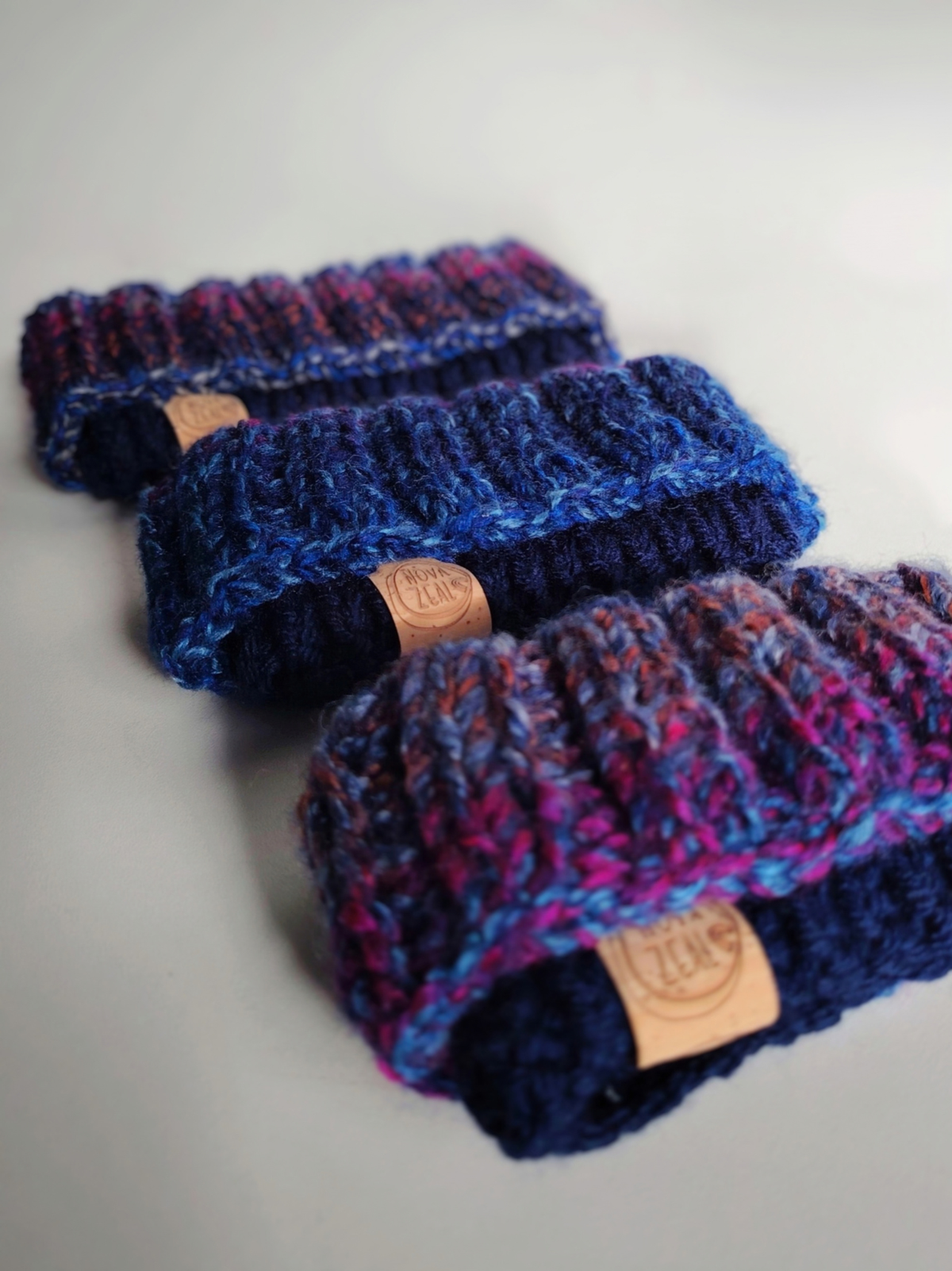 Three handmade Nova Zeal Night Sky dog snoods arranged on a light background, featuring navy knitted ribbing, blue, purple and pink yarn, and branded leather labels.