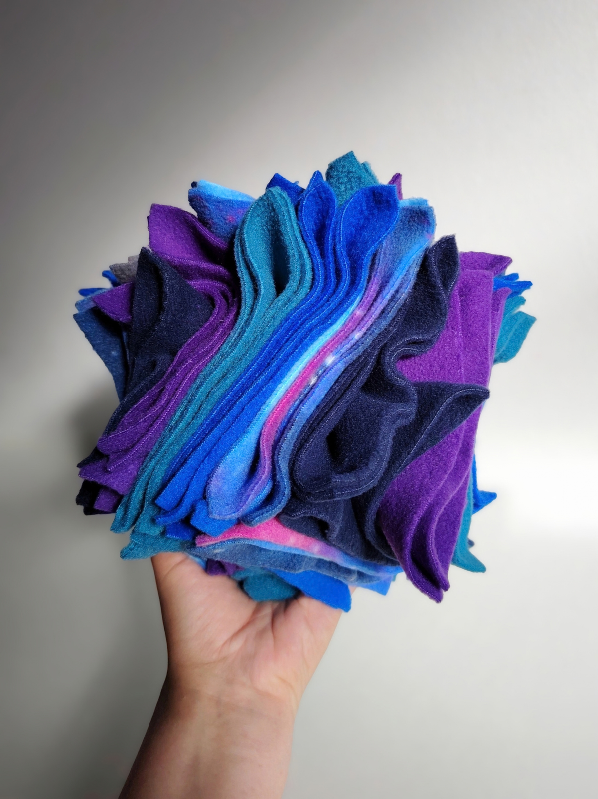 Hand holding a large Nova Zeal Night Sky Scruffle Ball, showing its size and layered folds of navy, royal blue, teal, purple and galaxy-patterned fleece.