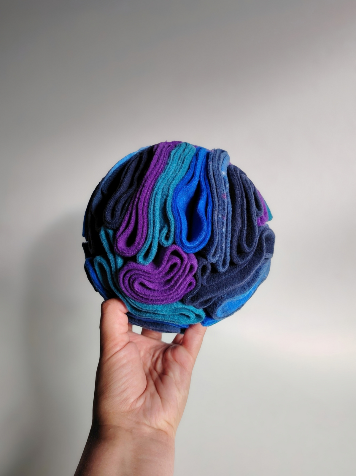 Medium Nova Zeal Night Sky Snuffle Ball held in one hand against a light background, showing its rounded shape and folded fleece in navy, royal blue, teal, purple and galaxy print.