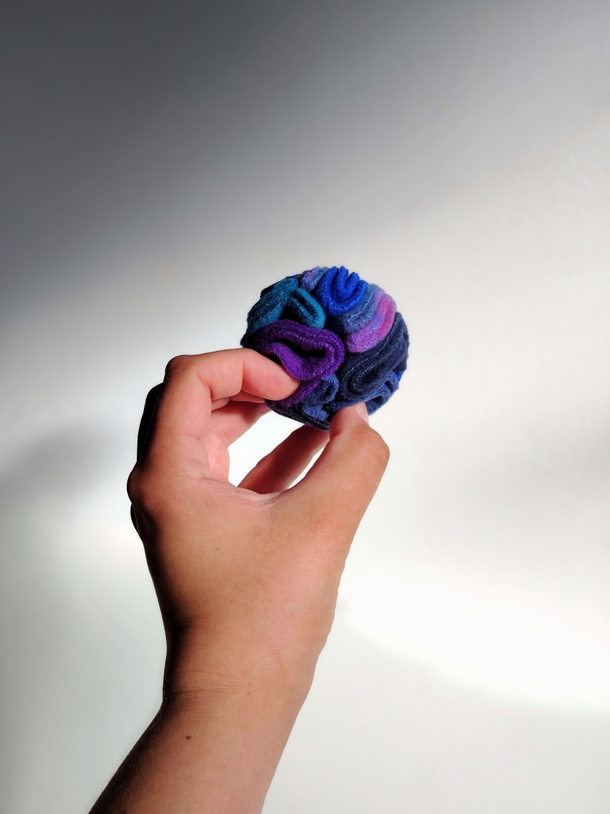 Hand holding a Nova Zeal Night Sky Pocket Snuffle Ball to show its compact size, featuring folded navy, royal blue, teal and purple fleece against a white background.