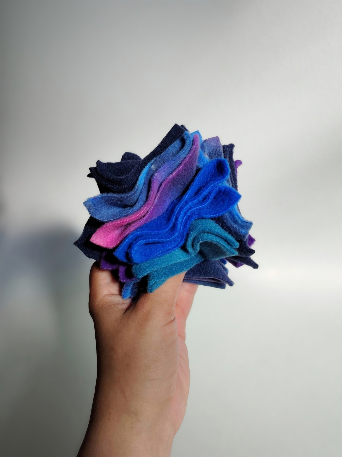 Hand holding a small Nova Zeal Night Sky Scruffle Ball, showing its compact size and folded layers of navy, royal blue, teal, purple and galaxy-patterned fleece.