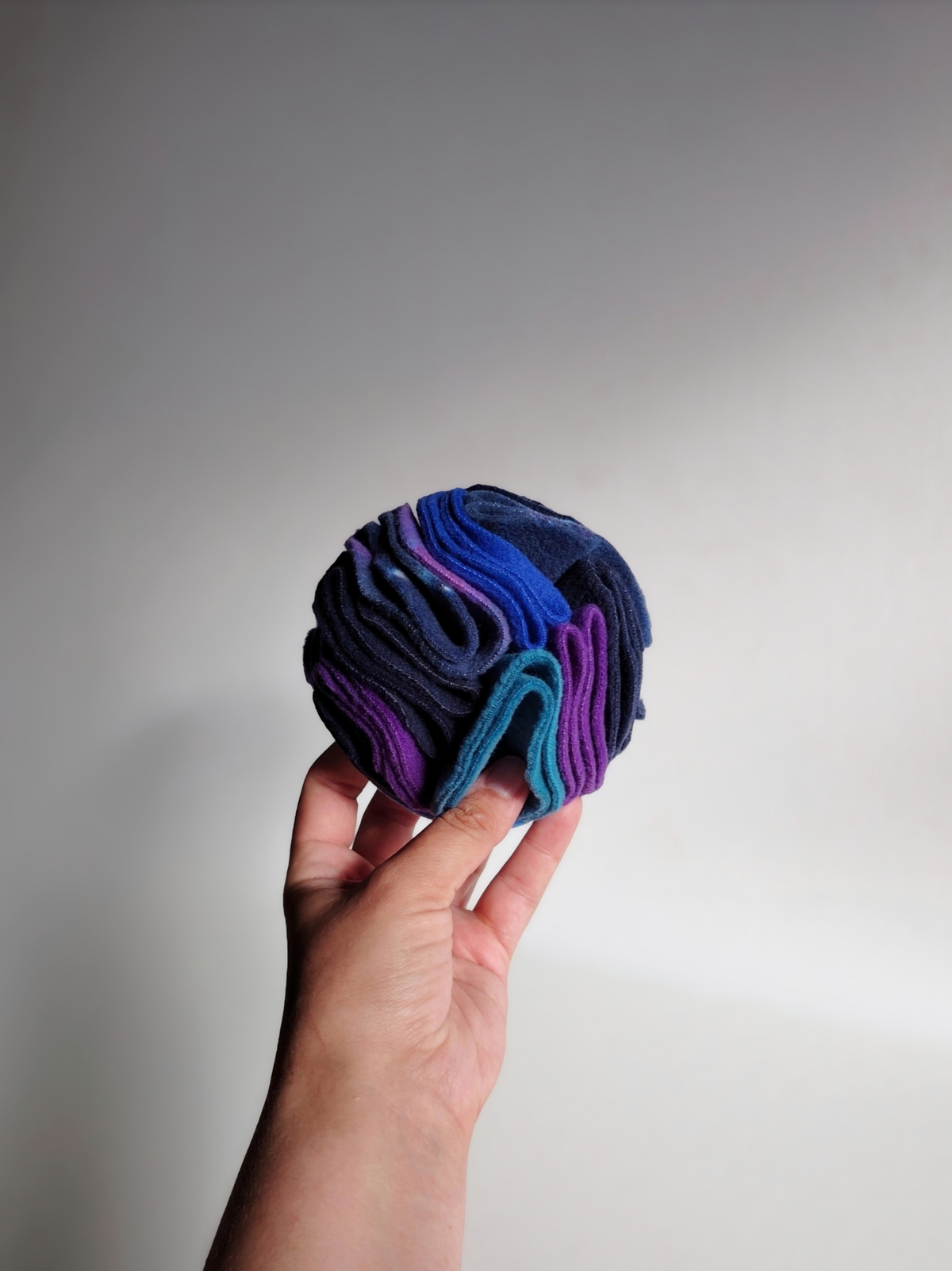 Hand holding a small Nova Zeal Night Sky Snuffle Ball against a light background, showing its rounded shape and layered navy, royal blue, teal and purple fleece folds.