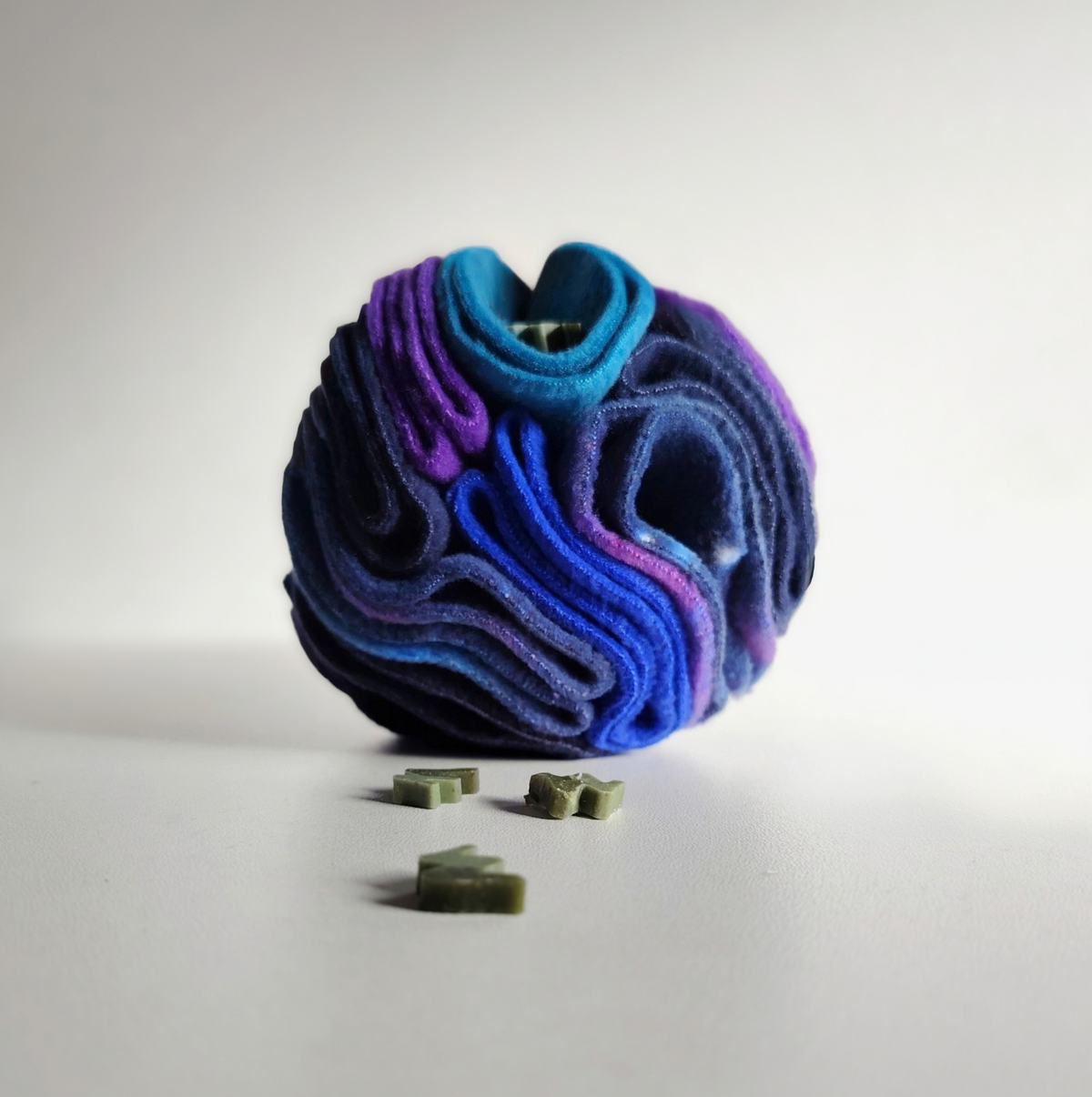 A Nova Zeal Small Snuffle Ball filled with treats in the layers of fleece in shades of blue, purple, and navy, photographed on a clean white studio background. 