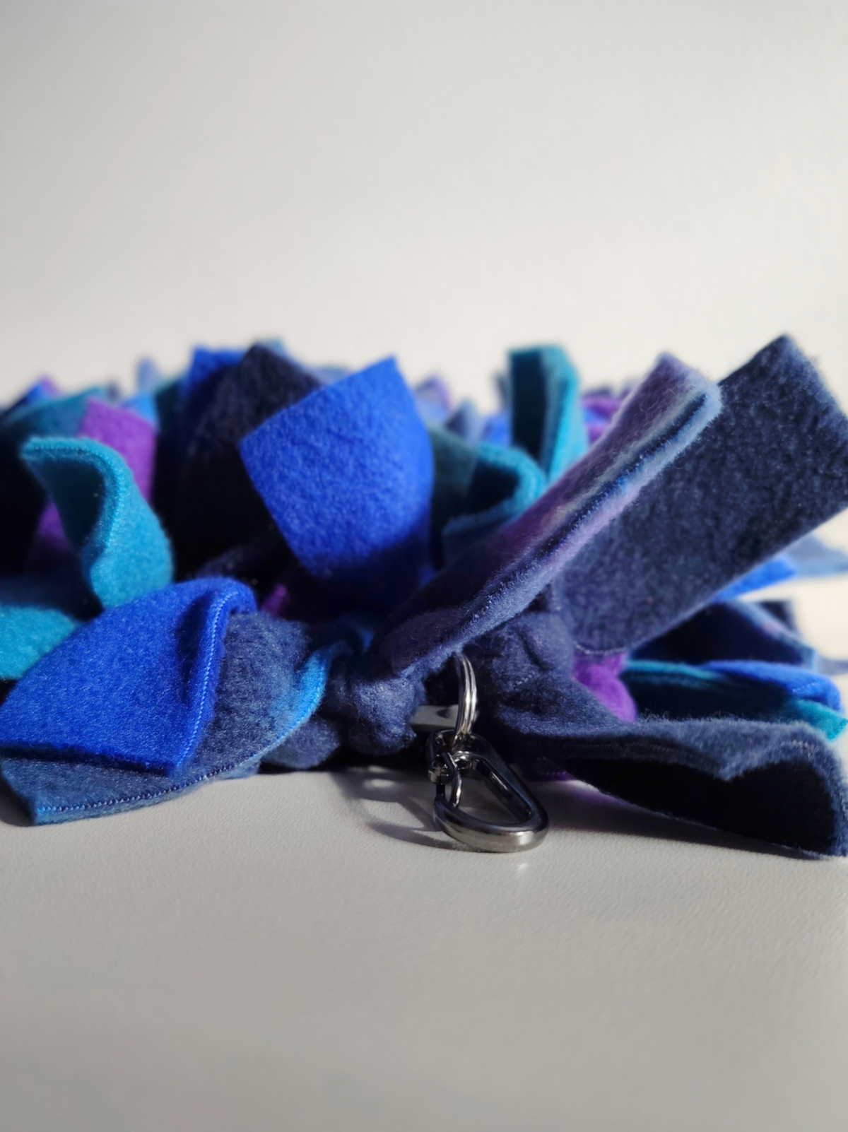 Close-up of the metal attachment clip on Nova Zeal's Night Sky Snuffle Mat, surrounded by navy, royal blue, teal and purple fleece strips.