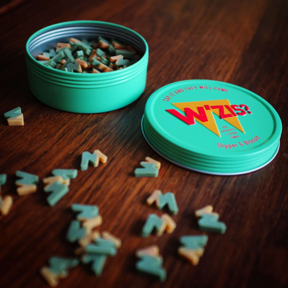 Open turquoise W'ZIS Lamp Post & Chips treat tin with small plant-based dog treats scattered on a wooden table.
