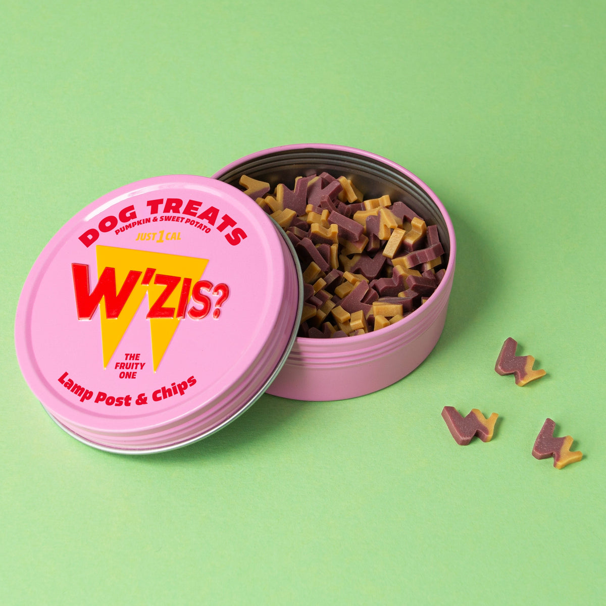 Pink W'ZIS Slipper & Biscuit plant-based dog treat tin open beside small treats on a green background.