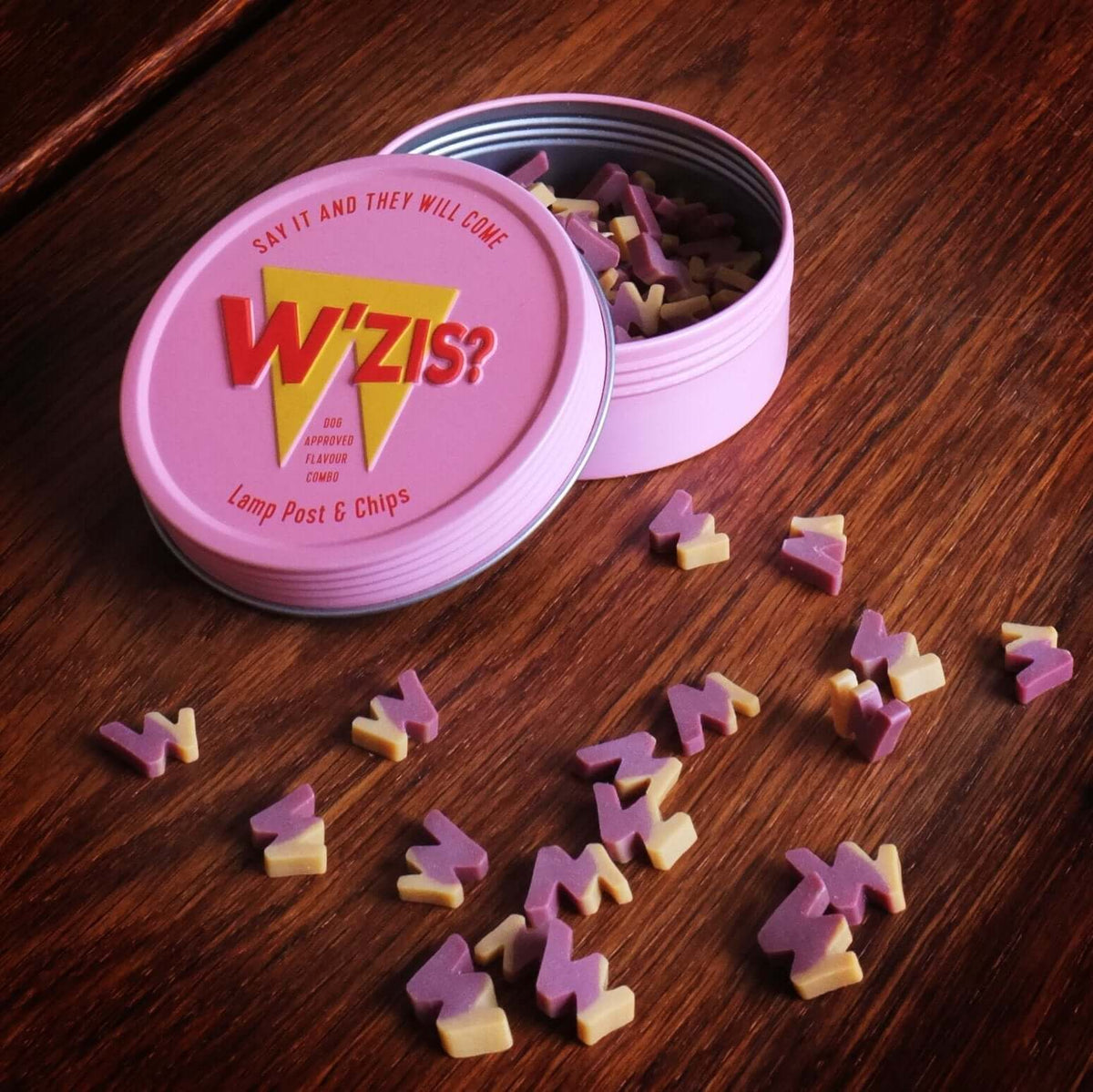 Open pink W'ZIS Slipper & Biscuit treat tin with small plant-based dog treats scattered on a wooden table.